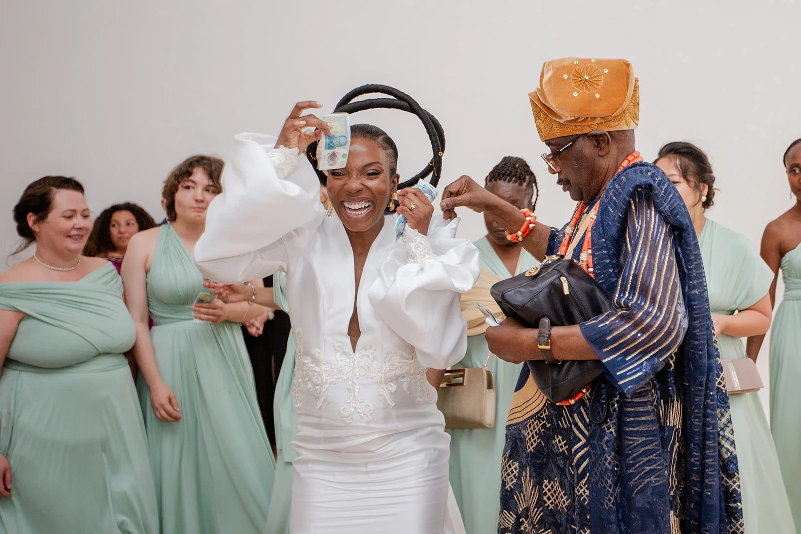 A joyful bride in a white gown with statement bell sleeves laughs ecstatically while money is sprayed during a traditional celebration.