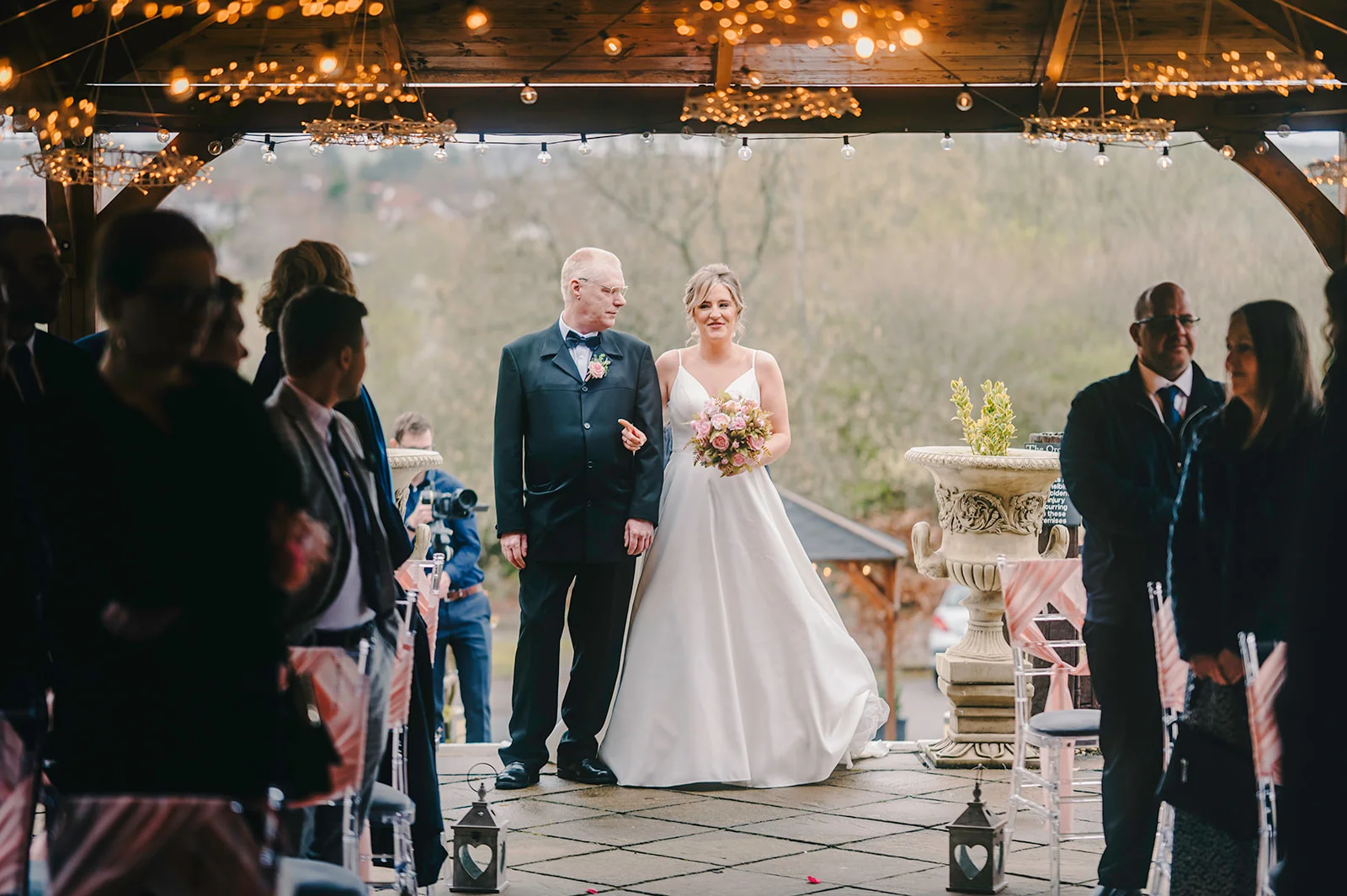A smiling bride holding a floral bouquet walks down the outdoor ceremony aisle linked with her father under a rustic wooden pavilion adorned with glowing fairy lights.