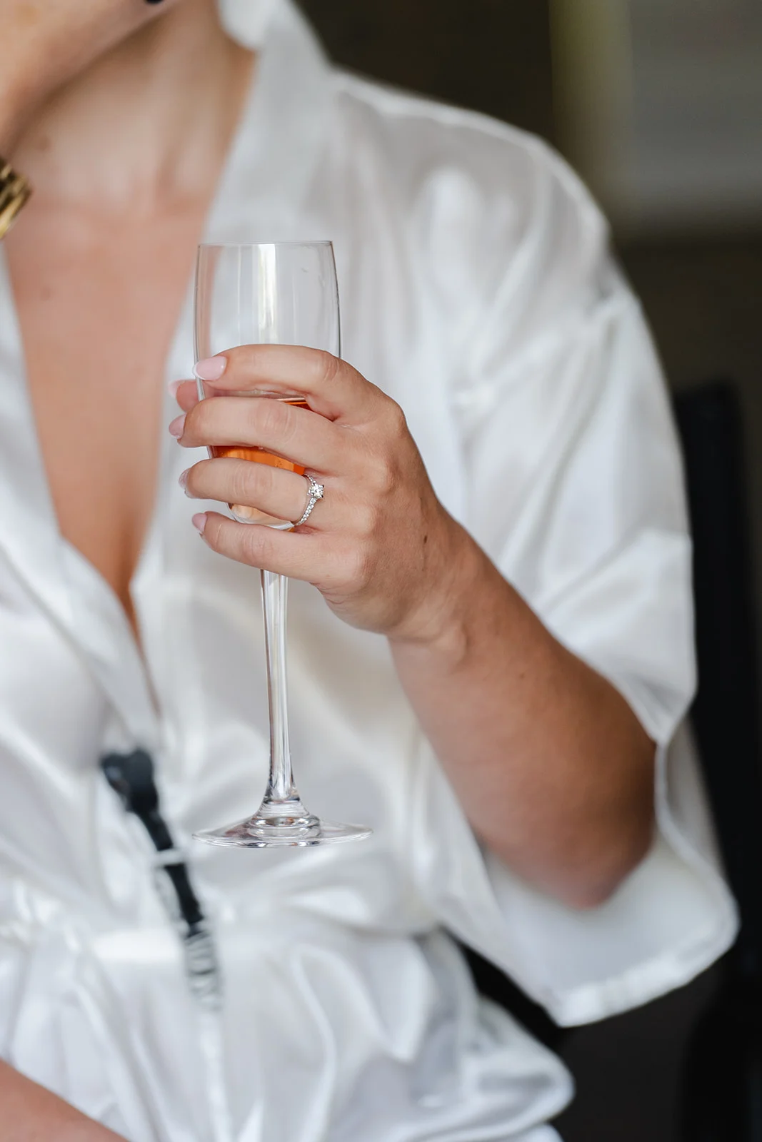 A bride wearing a white satin robe holds a champagne flute while showcasing her diamond engagement ring during wedding morning preparations.