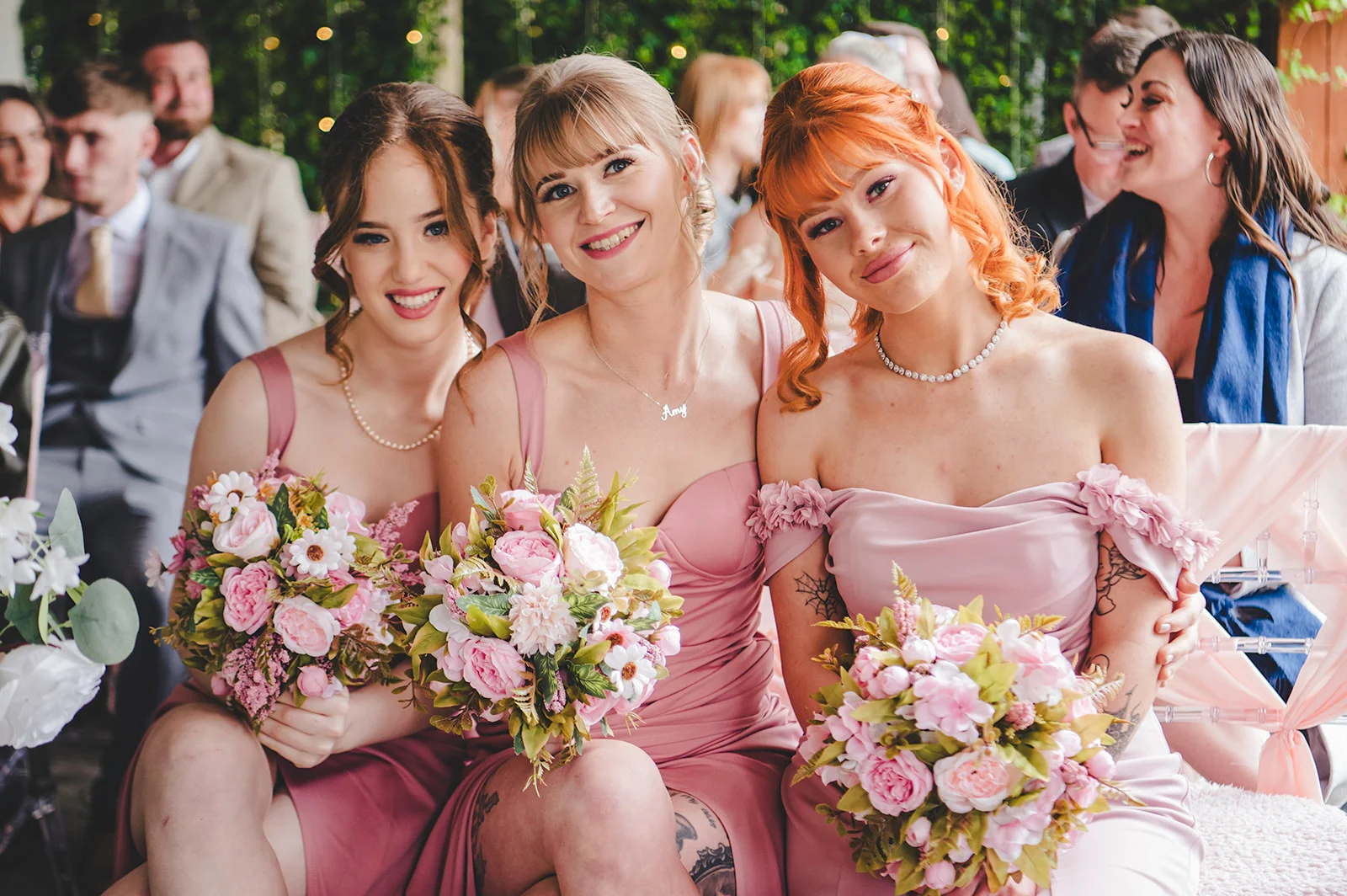 Three smiling bridesmaids in matching pink dresses hold lovely floral bouquets featuring pink roses during the wedding ceremony.