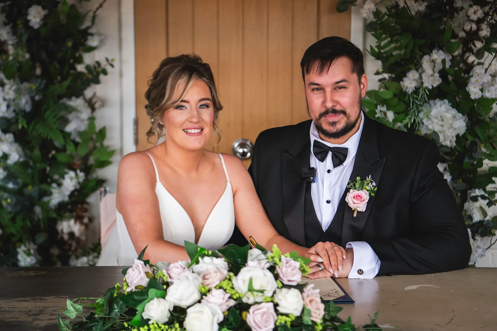 A smiling bride and groom sit side by side at a wooden table during the signing of the register, framed by lush floral arrangements of white and blush roses.