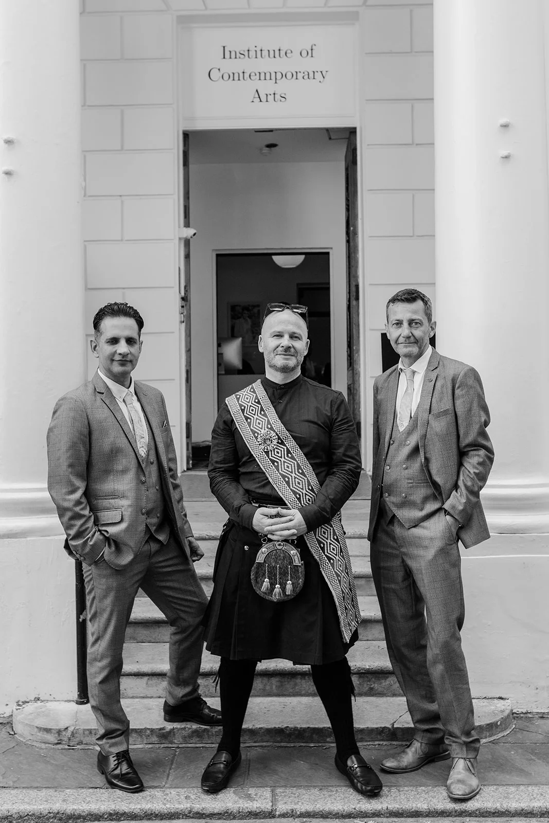 Three gentlemen pose together on the stone steps outside the Institute of Contemporary Arts in London, with the central figure wearing a traditional Scottish kilt and tartan sash flanked by two groomsmen in grey three-piece suits.