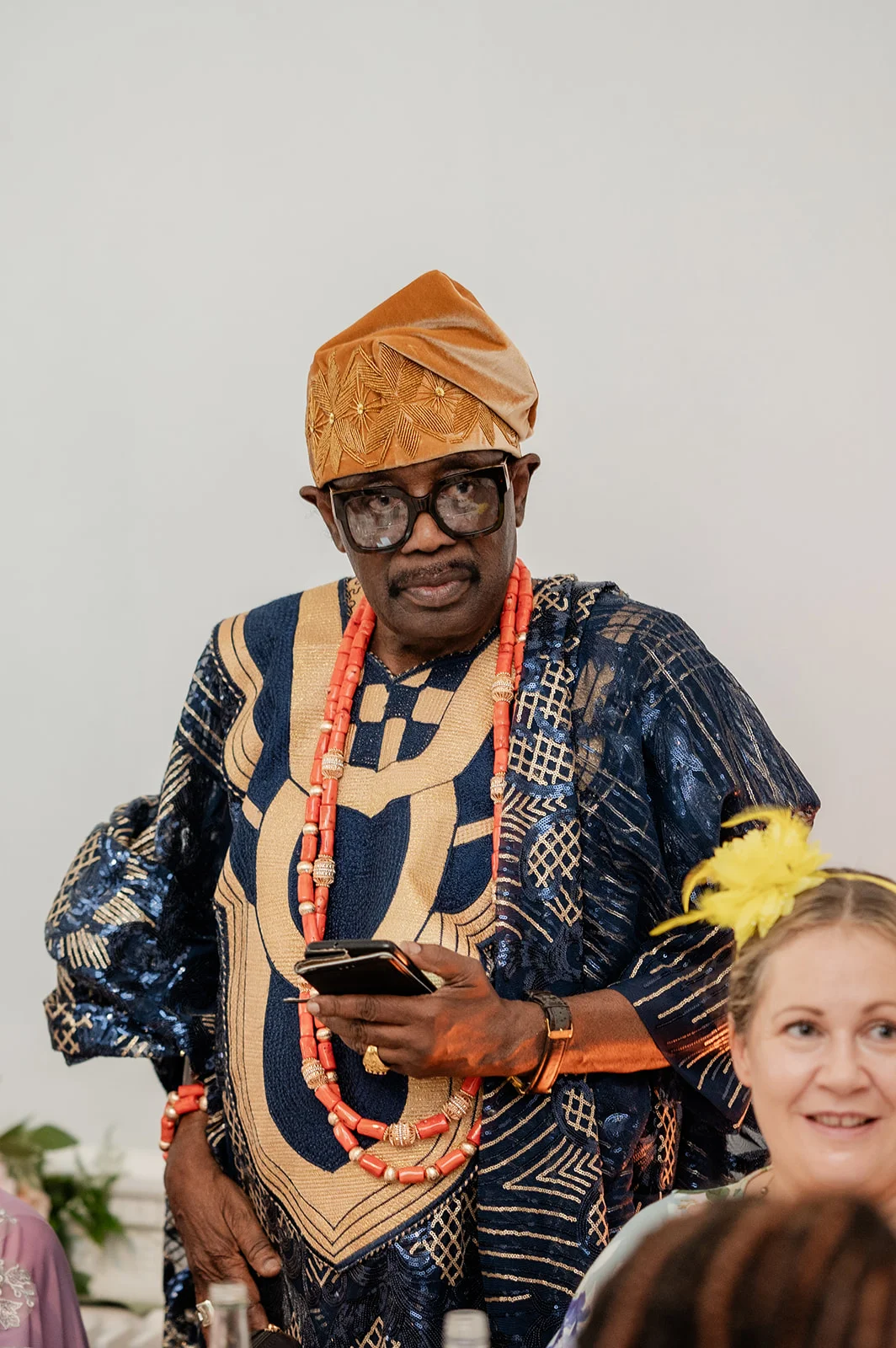 A guest at a wedding reception wears traditional Nigerian attire including a patterned blue tunic, coral bead necklaces, and a matching cap.