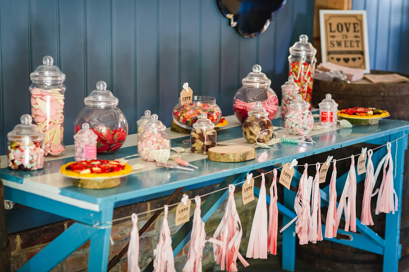 A colourful pick and mix sweet station is displayed on a blue wooden table adorned with pink tassel garlands and glass apothecary jars filled with retro sweets.