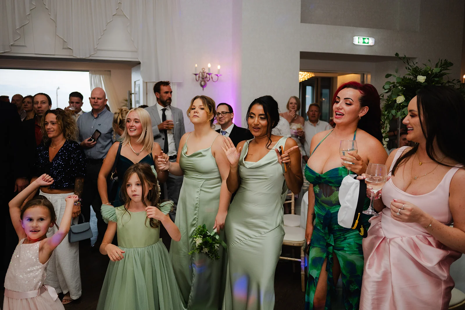 A lively group of wedding guests and bridesmaids in sage and pink dresses mingle and celebrate inside a brightly lit venue.