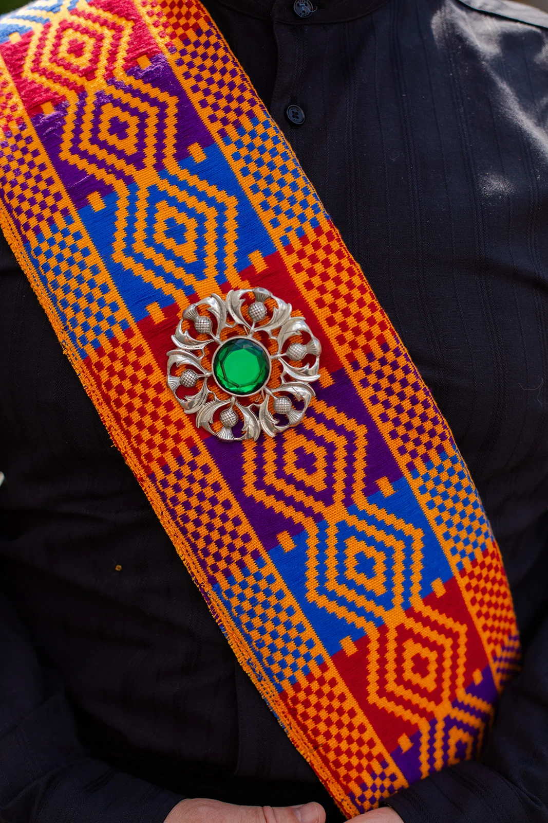 A close-up view of a vibrant patterned tartan sash worn across a dark shirt, secured with an intricate silver brooch featuring a bright green gemstone.