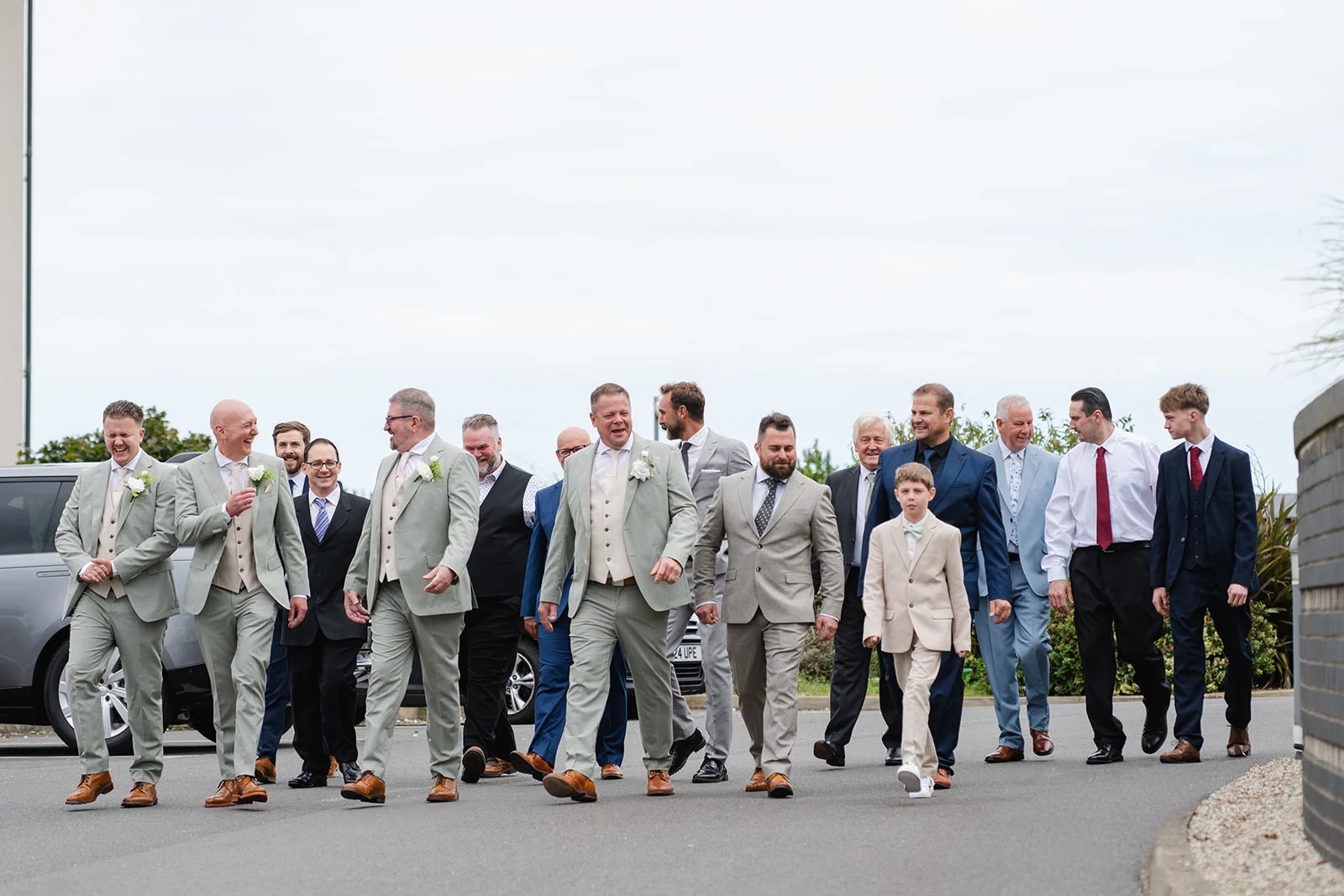 A cheerful group of groomsmen and wedding guests in suits walk outdoors, laughing and talking together.