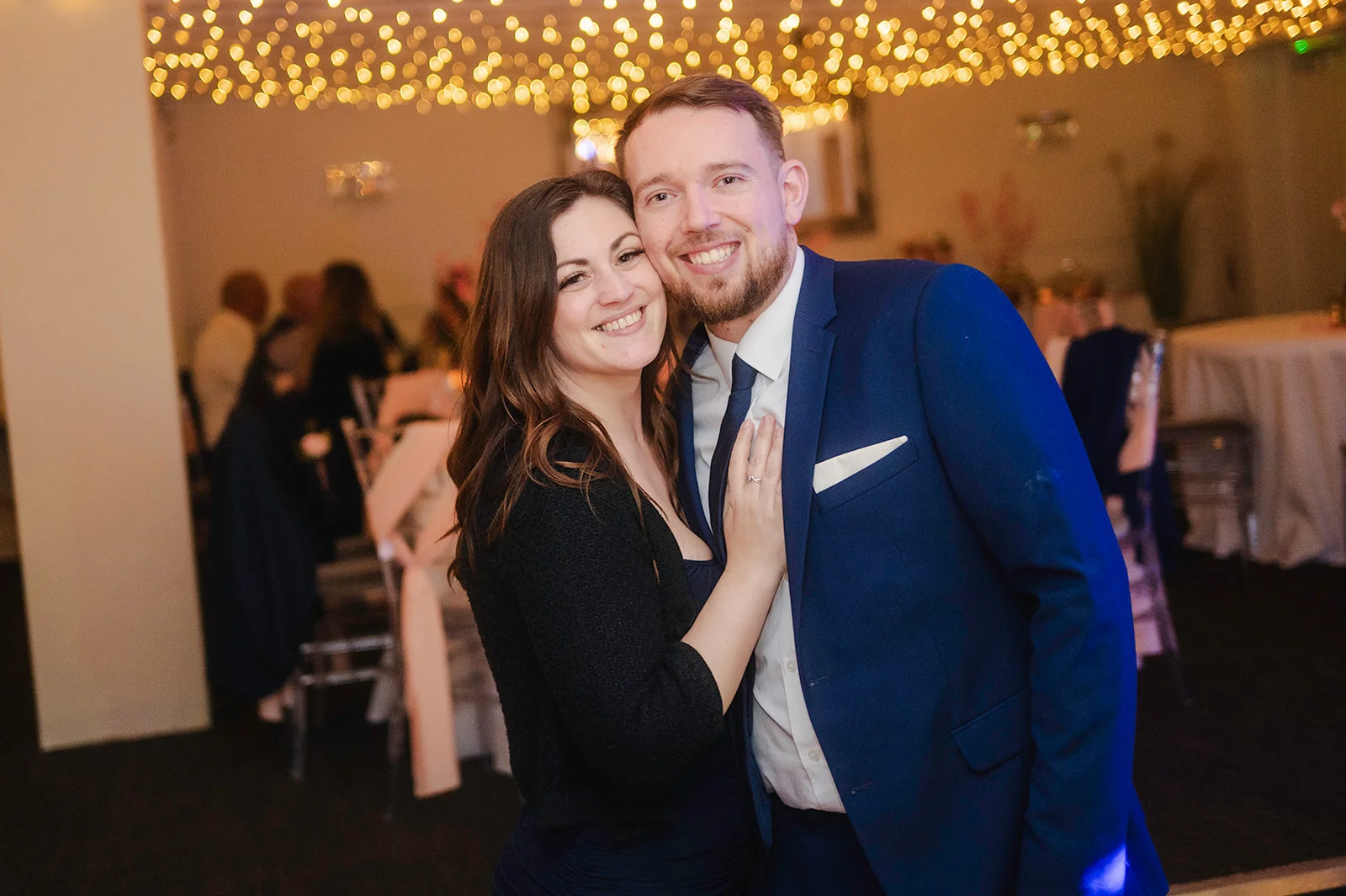 A smiling couple poses together during an evening wedding reception under a warm canopy of fairy lights.