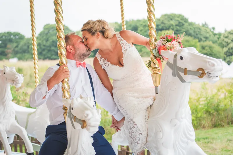 Newlyweds share a romantic kiss while sitting on white carousel horses at an outdoor festival-themed wedding celebration.