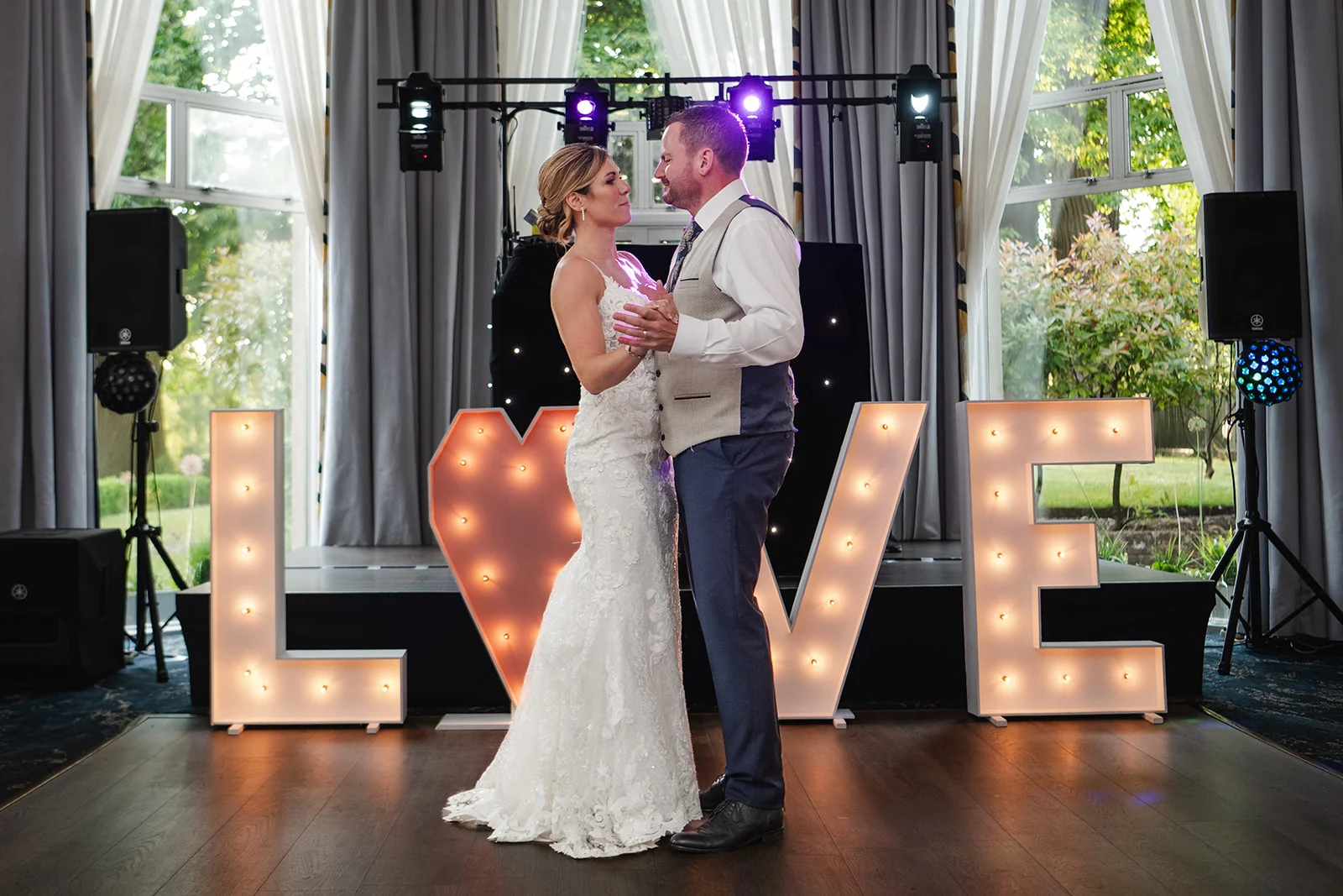 The newlywed couple share an intimate first dance on a wooden dance floor in front of illuminated light up love letters inside a brightly lit venue.
