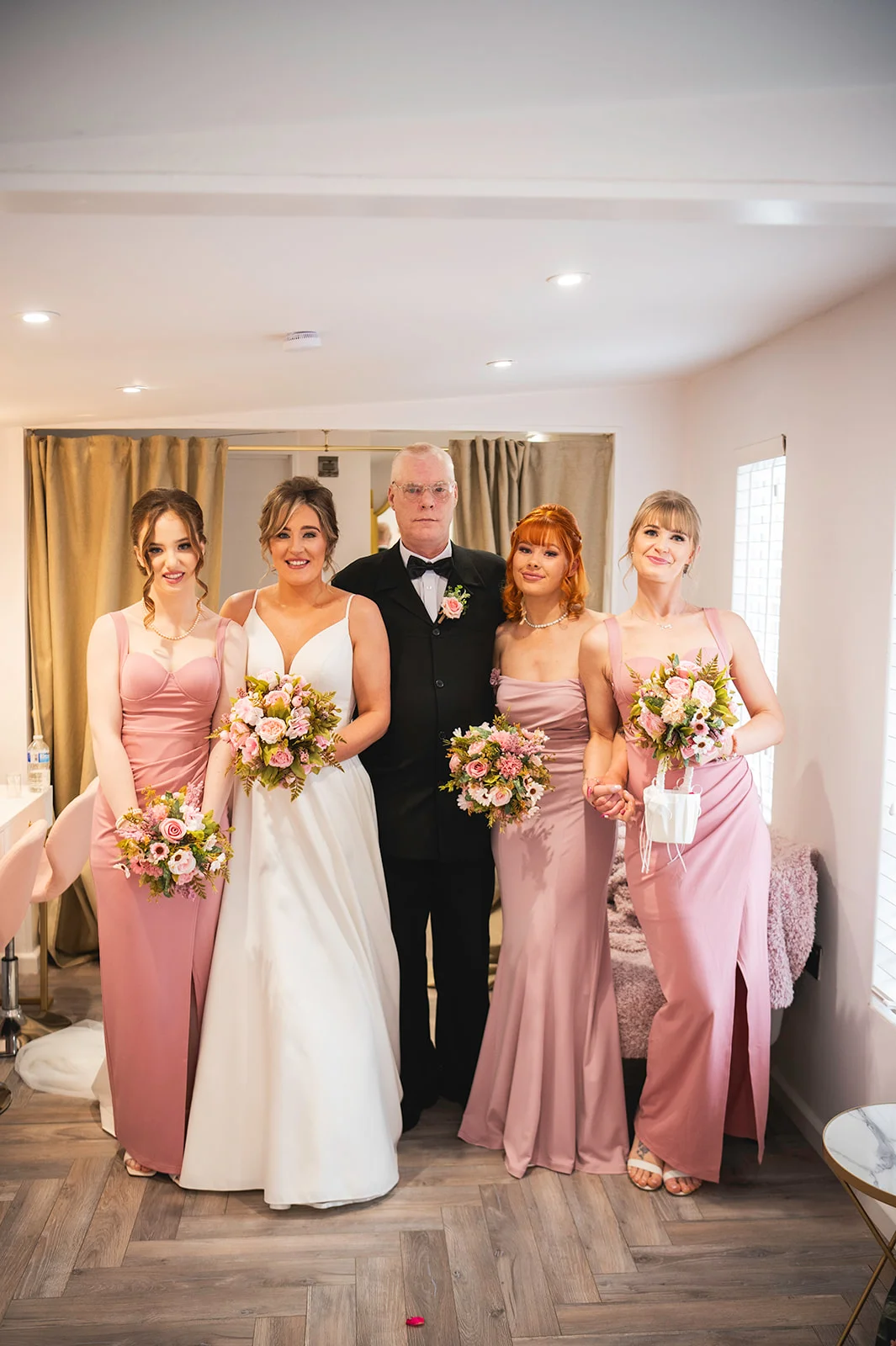 A bride in a white gown and three bridesmaids in pink dresses pose with a man in a black suit holding bouquets inside a bright room.