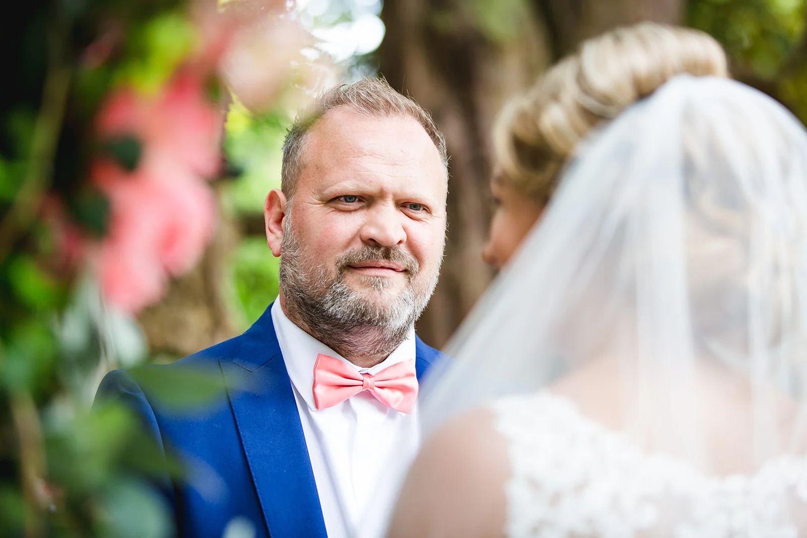An emotional groom in a blue suit and pink bow tie looks lovingly at his bride during an outdoor ceremony, framed by foliage.