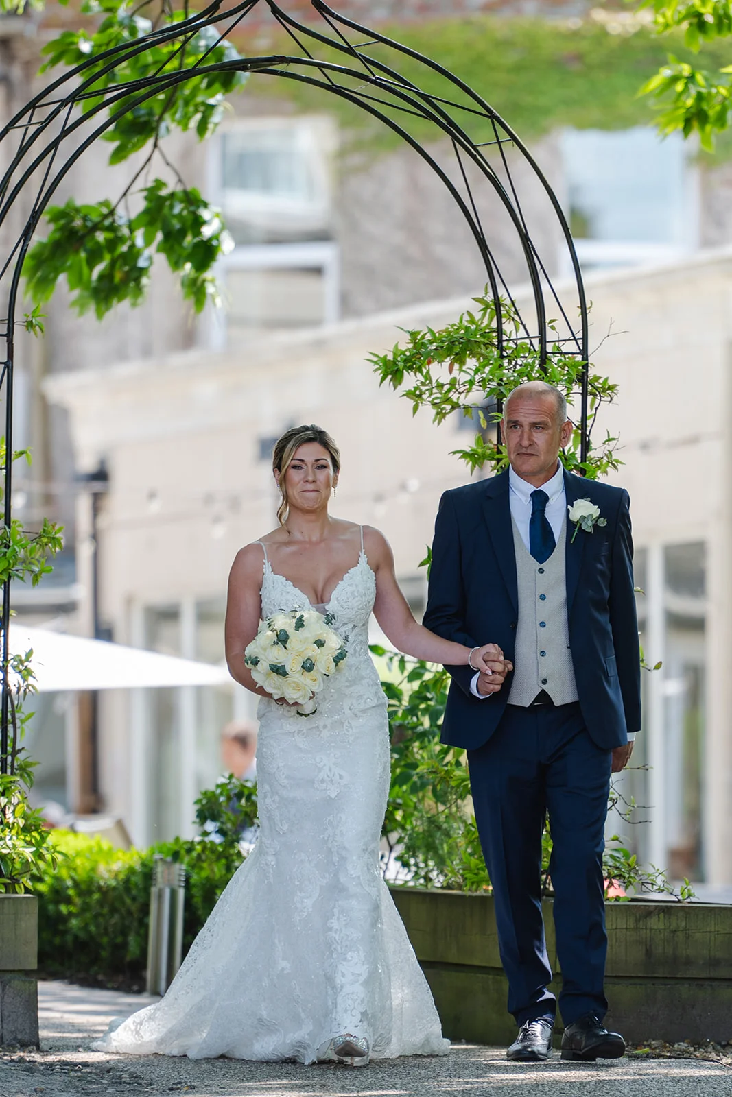 A bride holding a white rose bouquet walks down an outdoor aisle holding hands with her father beneath a metal garden archway.