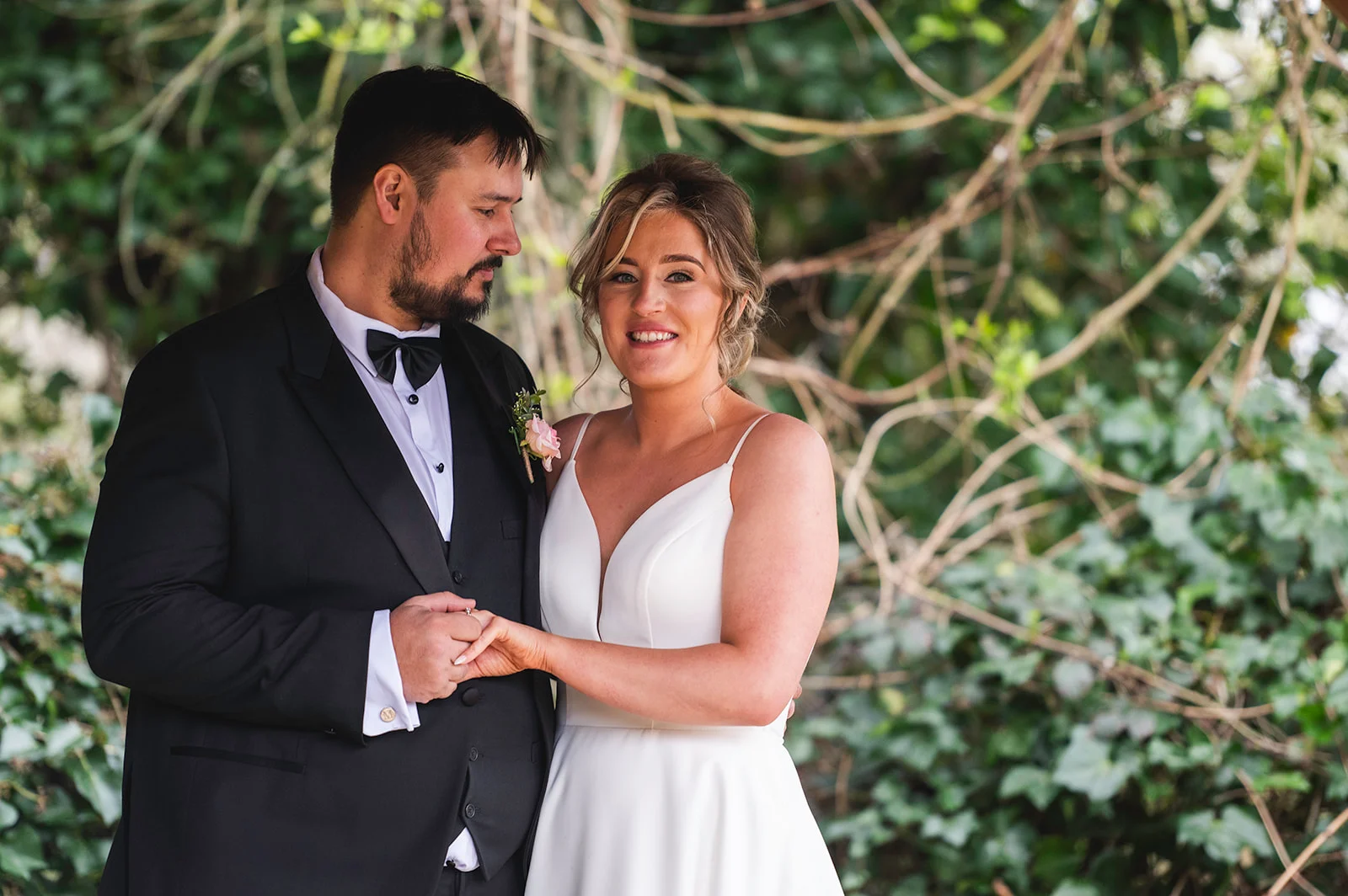 A newlywed couple stands closely together outdoors, holding hands while framed by lush green foliage and tree branches.