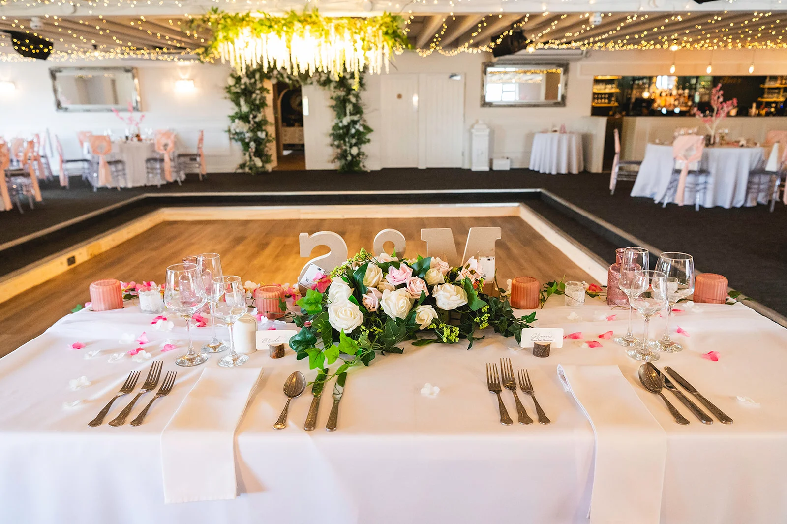 A beautifully styled sweetheart table featuring white linens, a lush floral centrepiece with white and pink roses, wine glasses, and place settings, set in a venue with a wooden dance floor and fairy light canopy.