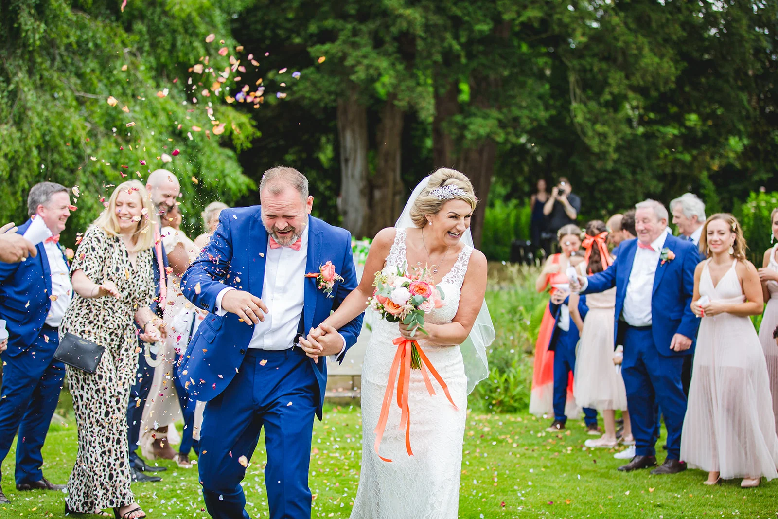 A joyful newlywed couple walks hand in hand through a shower of colourful confetti thrown by wedding guests in a lush garden setting.