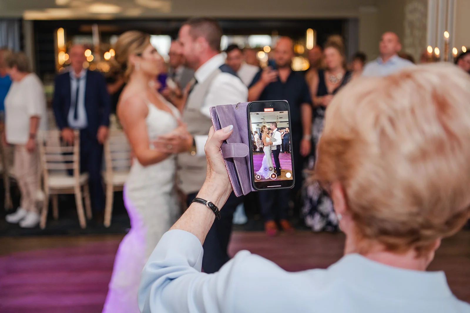 A guest records video on a smartphone of the bride and groom sharing their first dance at the wedding reception.