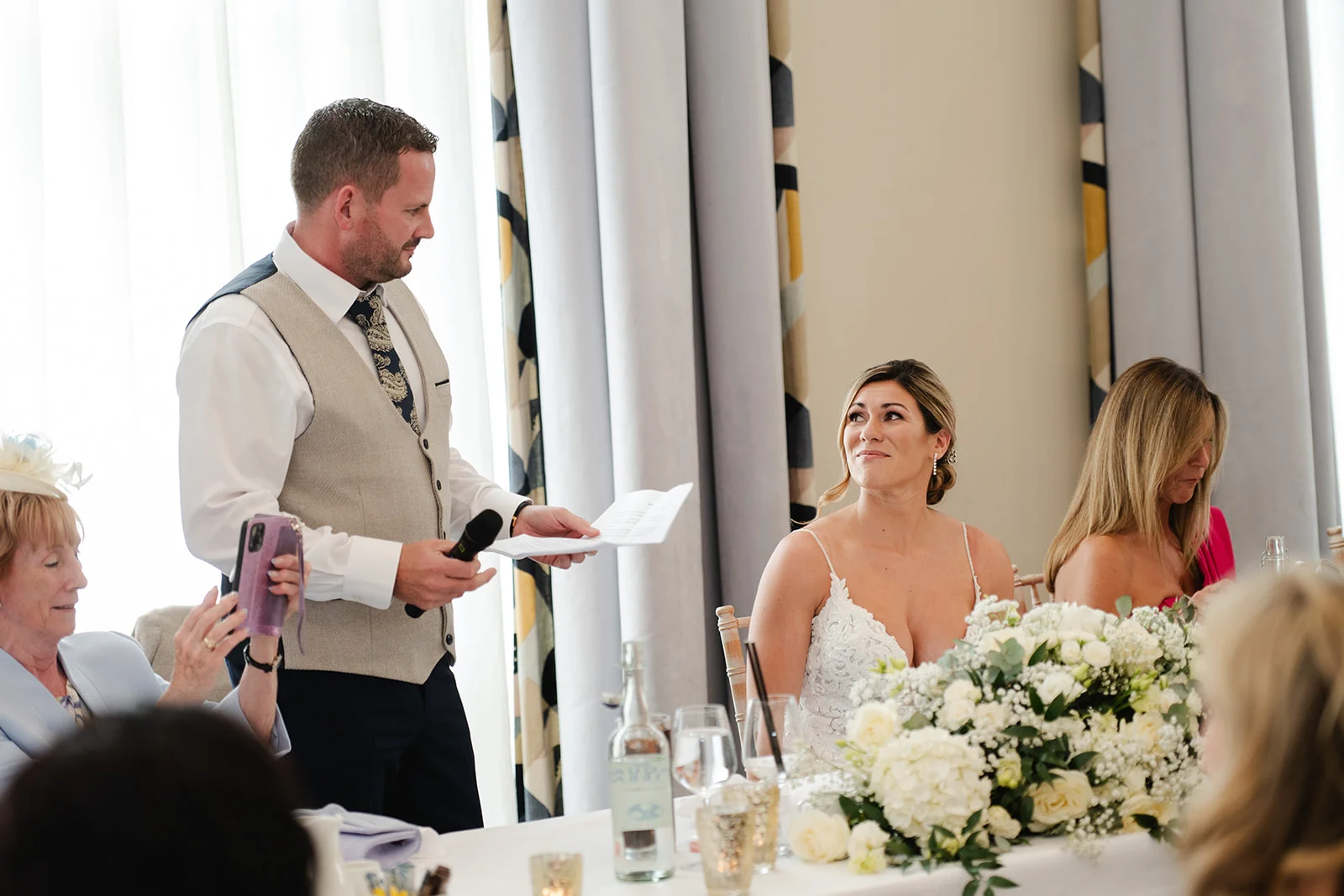 A groom delivers a heartfelt speech holding notes and a microphone, while the bride listens emotionally with a smile next to a floral tablescape.