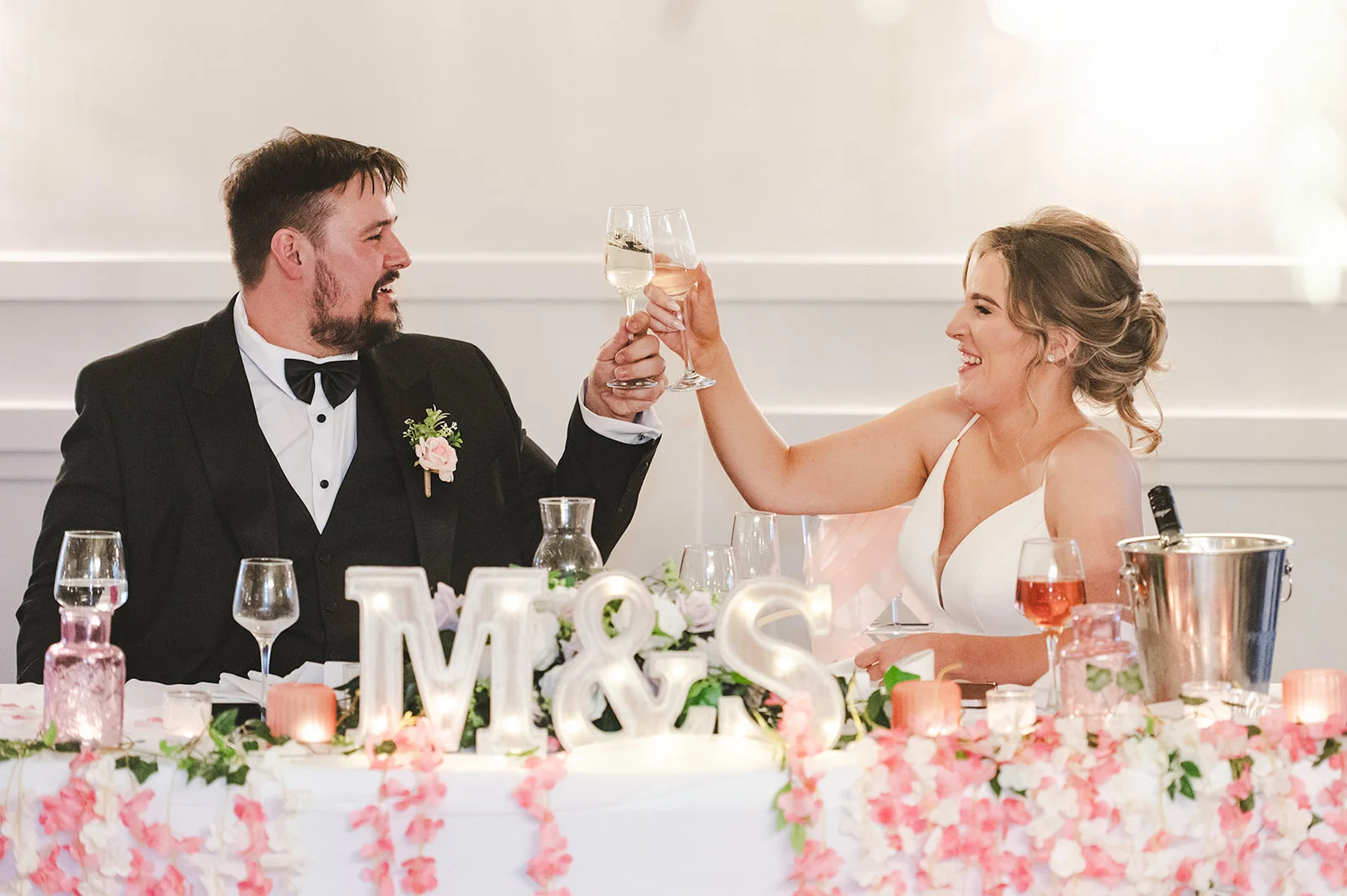 A smiling bride and groom in formal attire raise their champagne glasses to toast during the wedding reception at the top table.