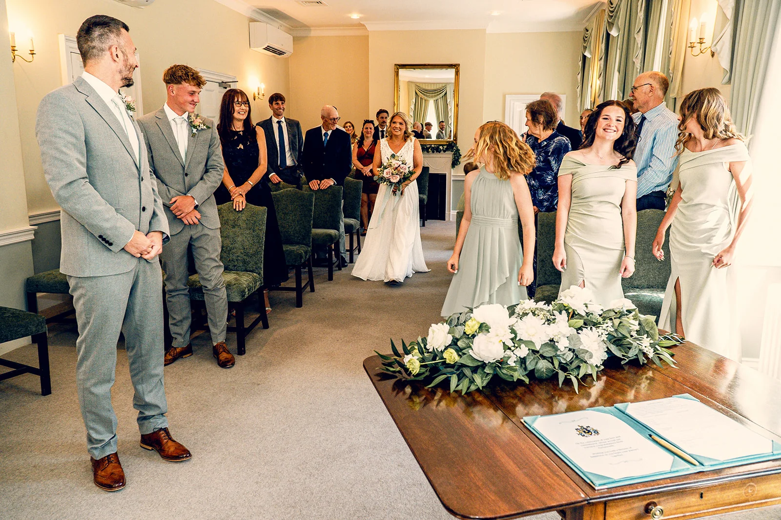 A smiling bride walks down the aisle holding a bouquet towards the groom and wedding party during an indoor civil ceremony.