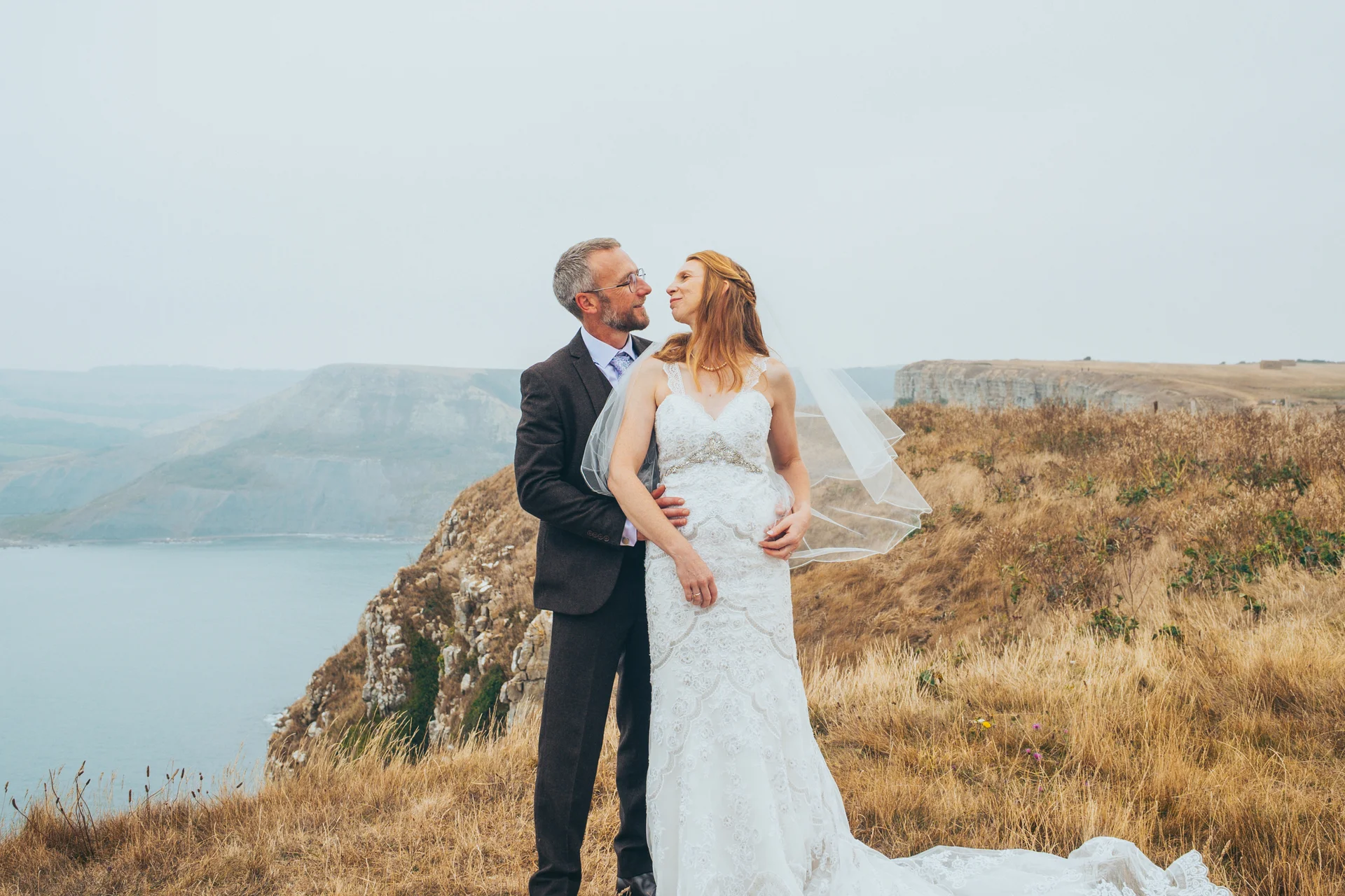 An older bride and groom gaze lovingly at each other during an outdoor portrait session on a dramatic clifftop overlooking the sea.