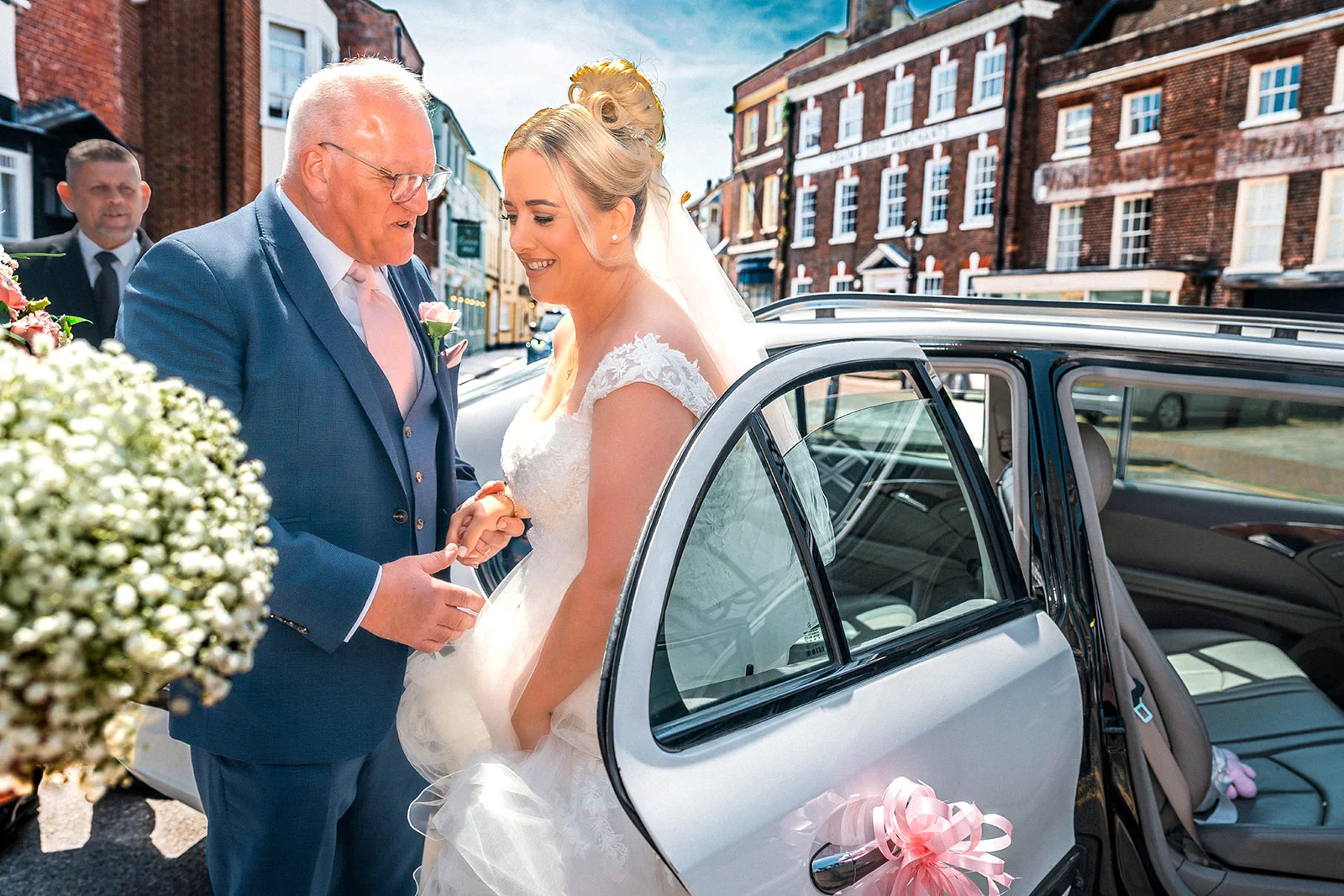 A smiling bride holding a bouquet gets ready to step out of a wedding car while sharing a tender moment with her father in a sunny town setting.