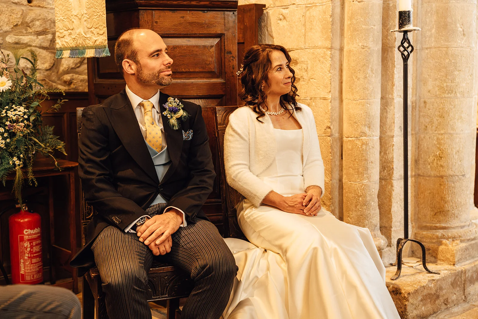 A bride and groom are seated together inside a stone church during their wedding ceremony, both looking attentively forward.