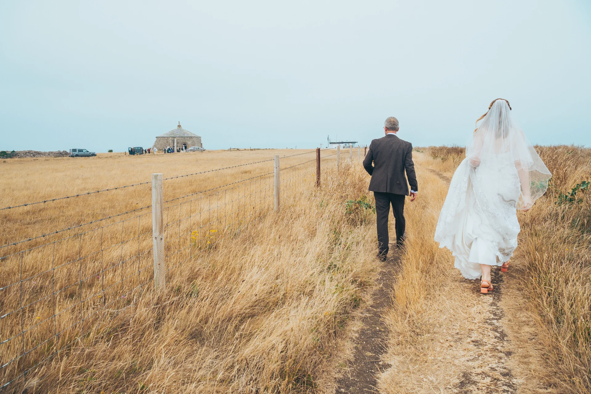 A bride and an older man in a brown suit walk along a grassy path beside a wire fence towards a remote stone chapel under an overcast sky.