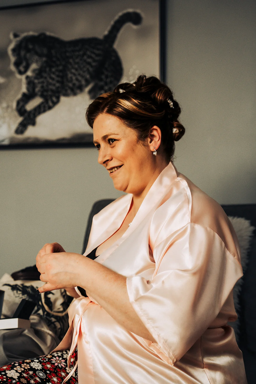 A mature bride wearing a pale pink satin getting-ready robe and an elegant updo smiles warmly while seated indoors.