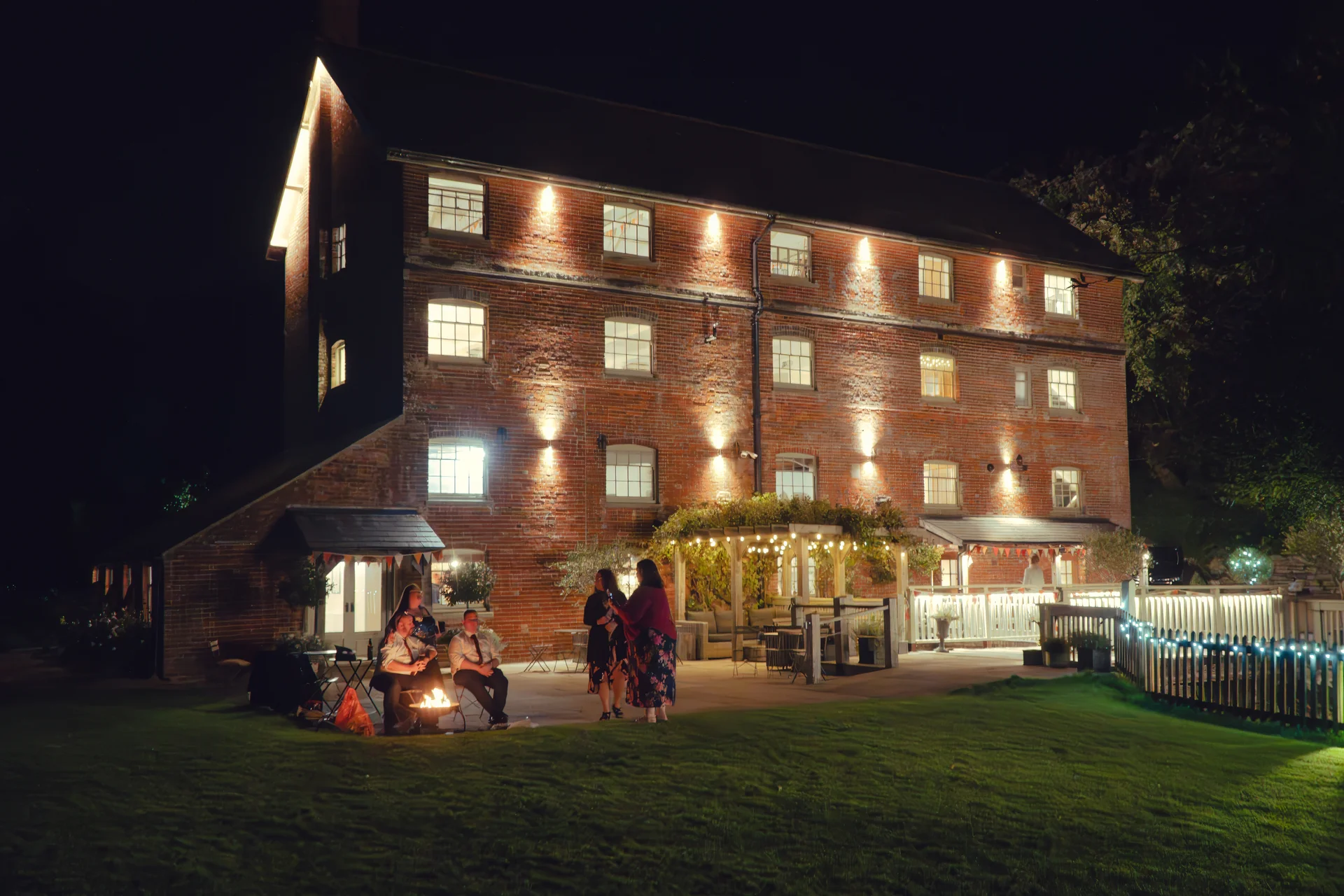 Guests enjoy a late-night gathering around a glowing firepit outside an illuminated multi-story brick wedding venue at night.