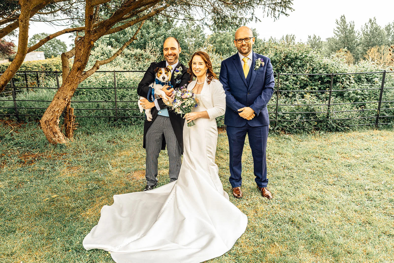 A smiling bride and groom pose outdoors in a lush garden setting, with the groom holding a small dog in a blue bow tie and a second man standing alongside them.