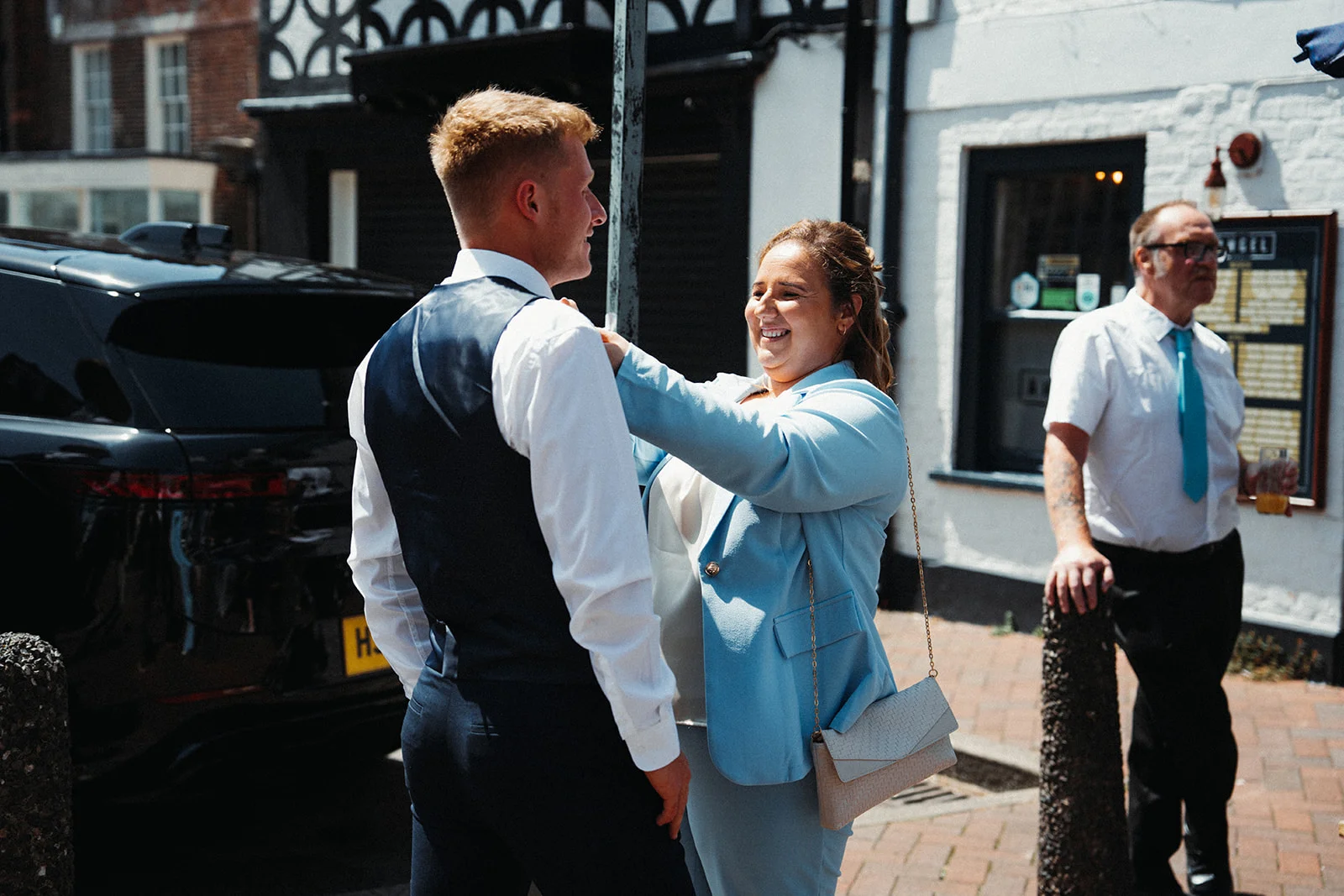 A smiling guest in a powder blue suit jacket helps adjust the lapel of a groom wearing a navy waistcoat outside a traditional British pub on a bright sunny day.