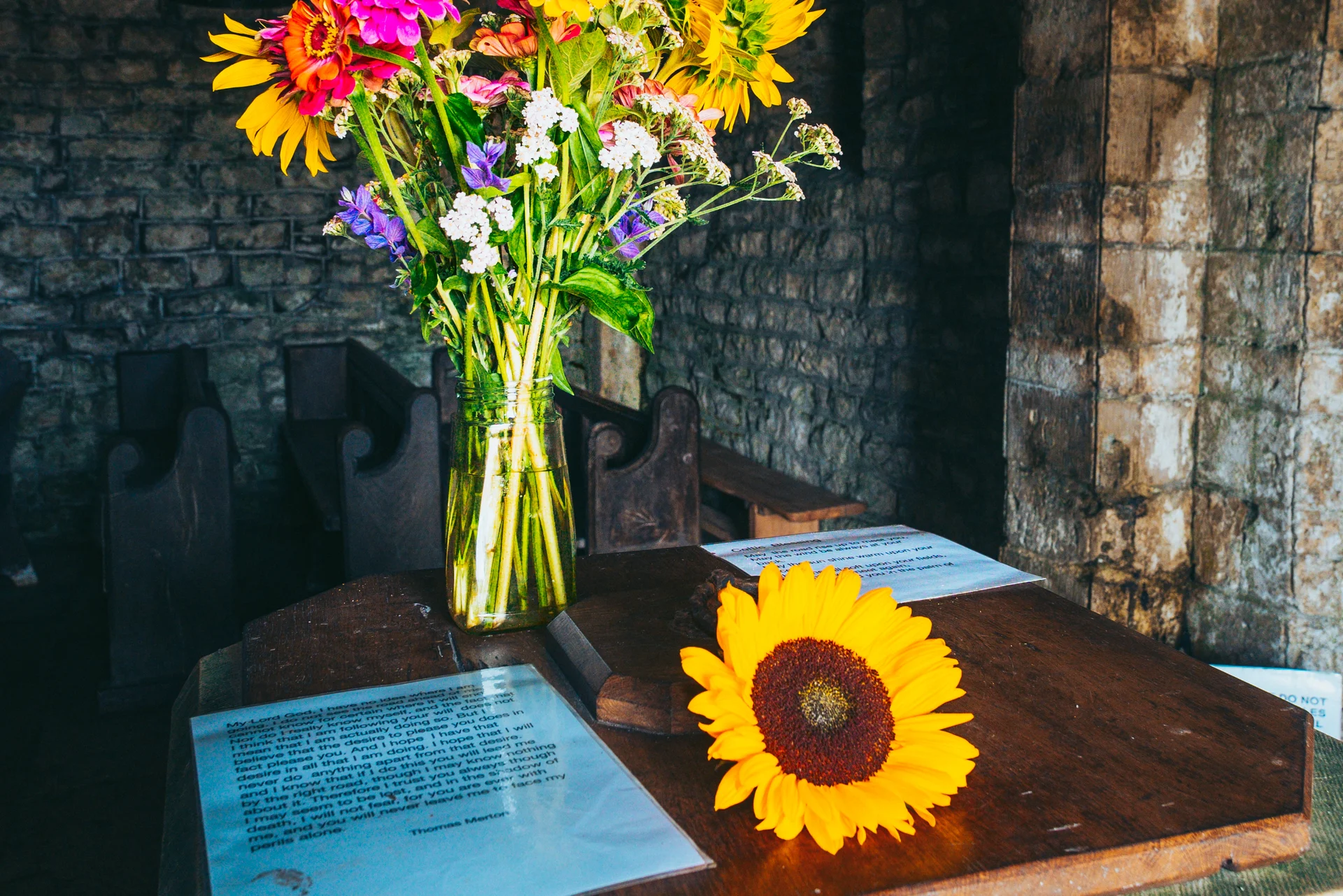 A vibrant bouquet of wildflowers and sunflowers in a glass mason jar sits on a dark wooden podium inside a stone church.