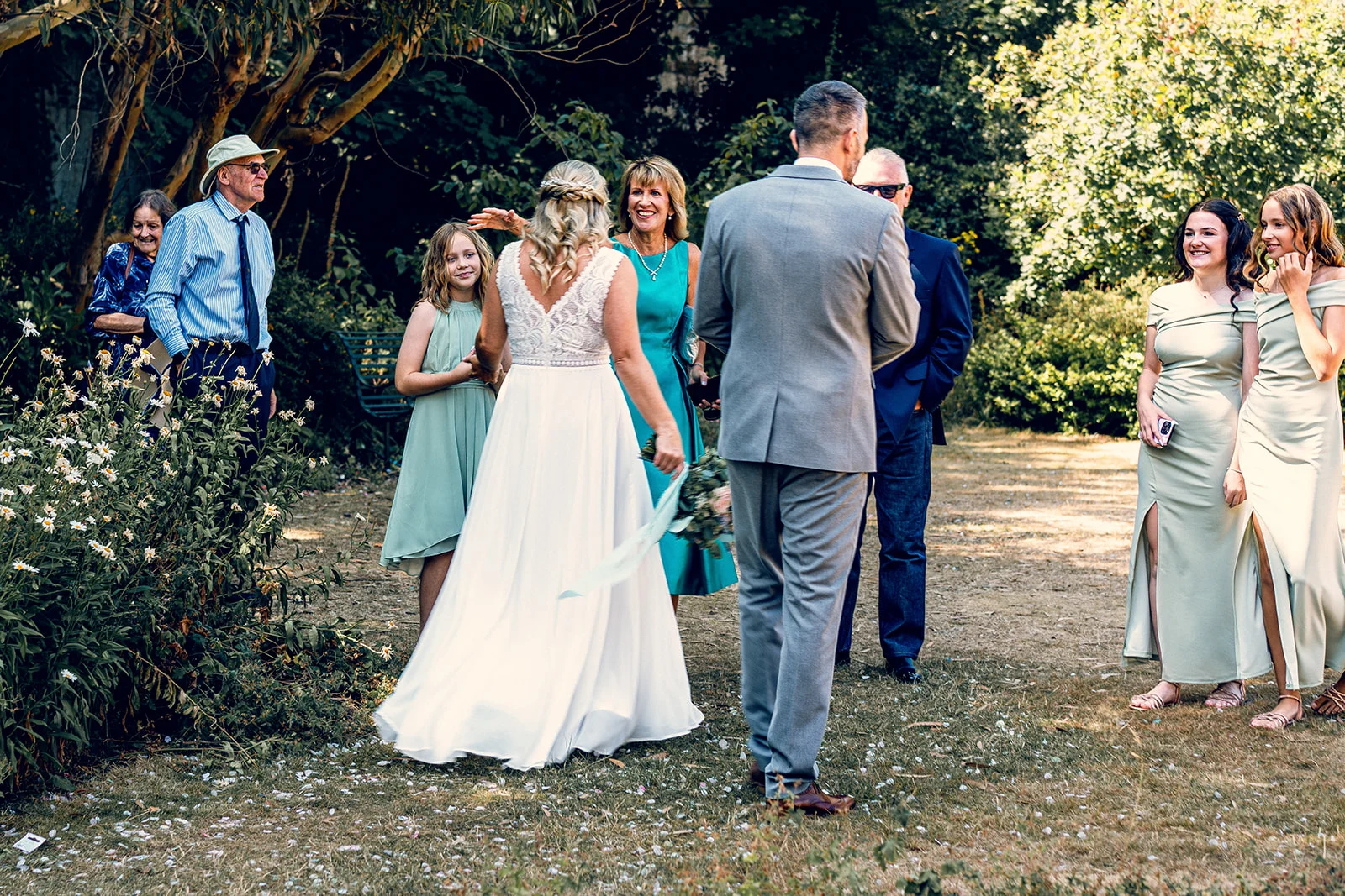 A bride in a lace-detailed a-line gown and a groom in a grey suit stand on a grassy lawn surrounded by family and bridesmaids in sage dresses during an outdoor garden wedding.
