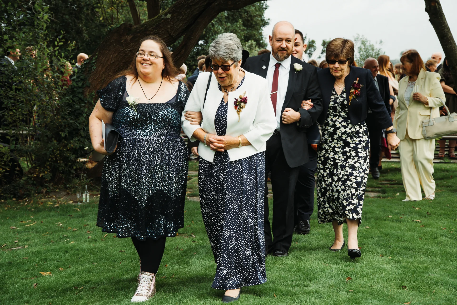 Family members and wedding guests happily walk together across a lush green lawn during an outdoor wedding celebration.