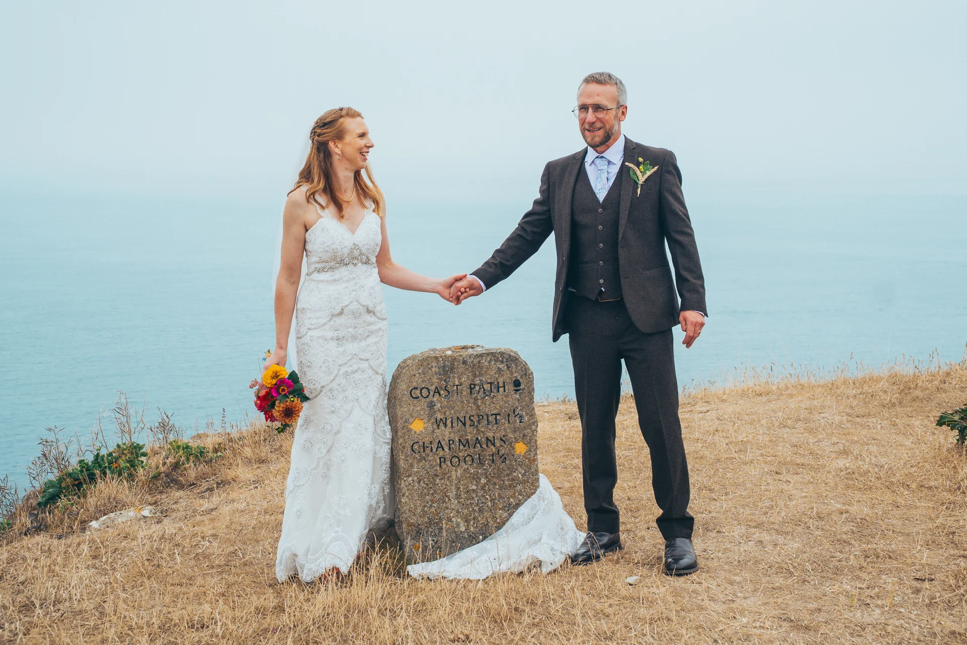 A newlywed couple holds hands by a stone coast path marker on a grassy clifftop overlooking the sea.