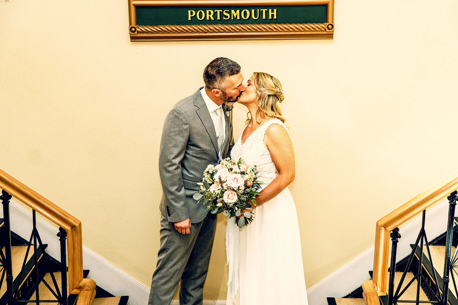 A newlywed couple shares a tender kiss beneath a Portsmouth sign framed by a sweeping staircase inside a venue.