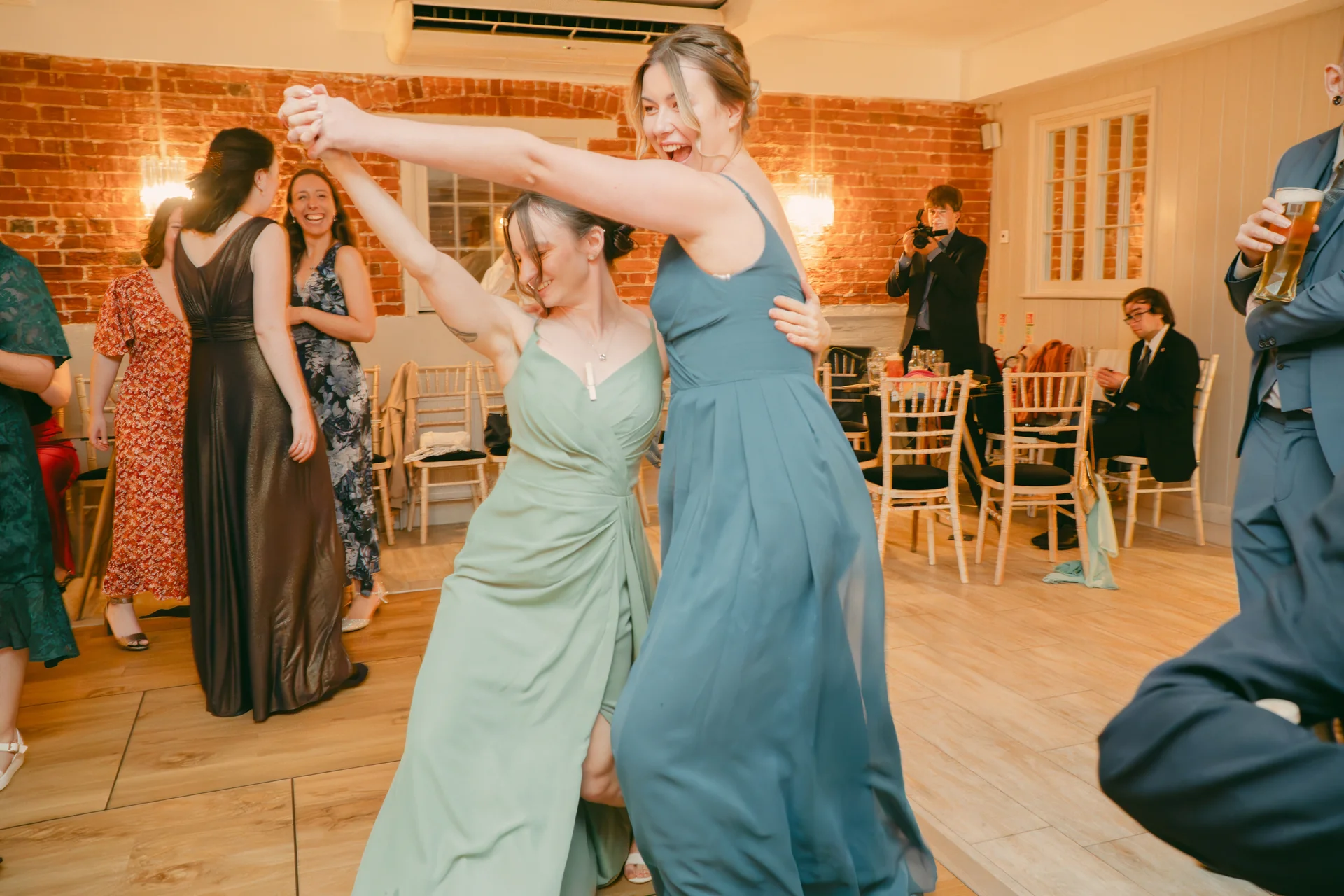 Two brides laughing and dancing joyfully together on a wooden dance floor in a rustic pub wedding venue with exposed brick walls.