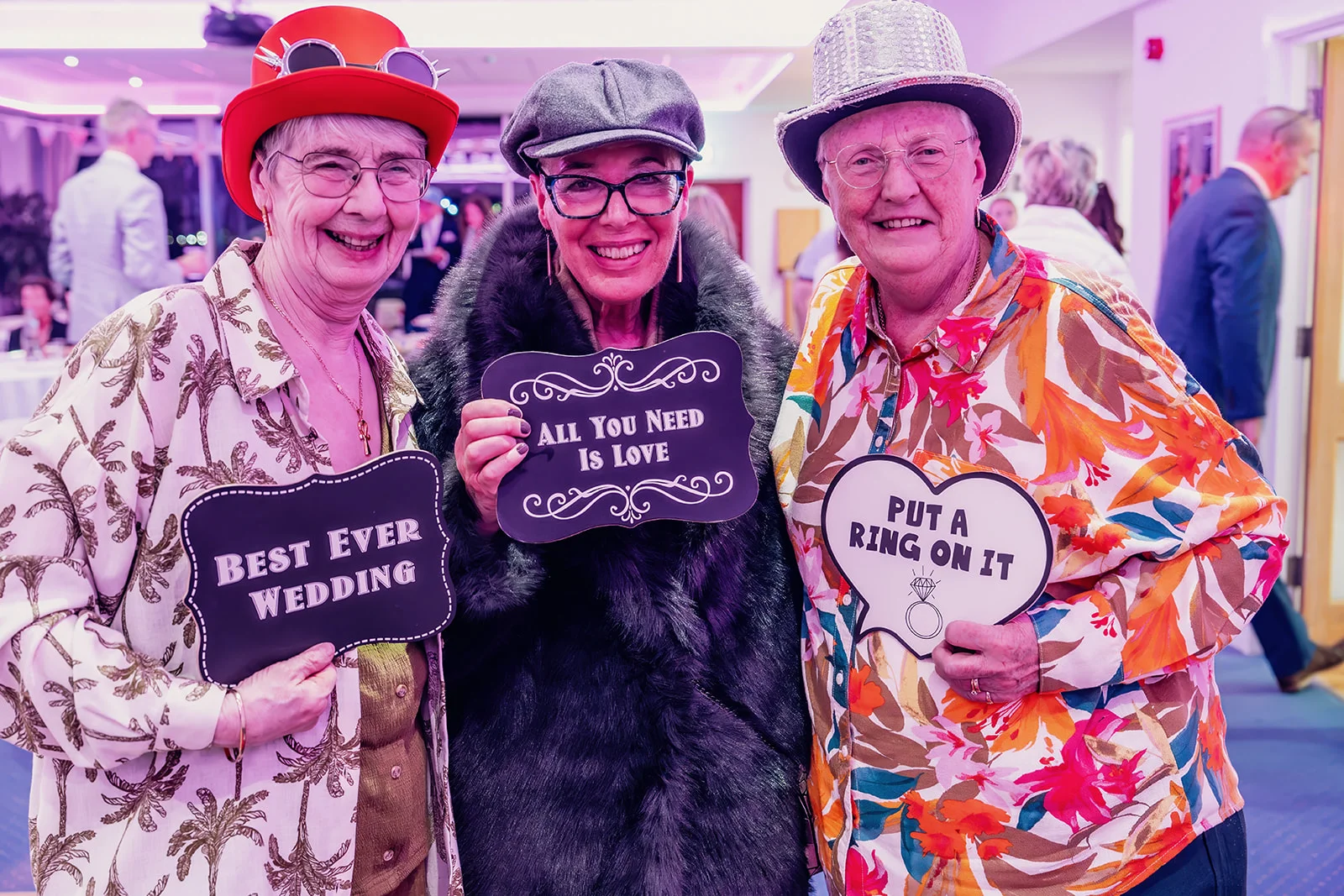 Three smiling older guests pose happily at a wedding reception while holding humorous photo booth signs.
