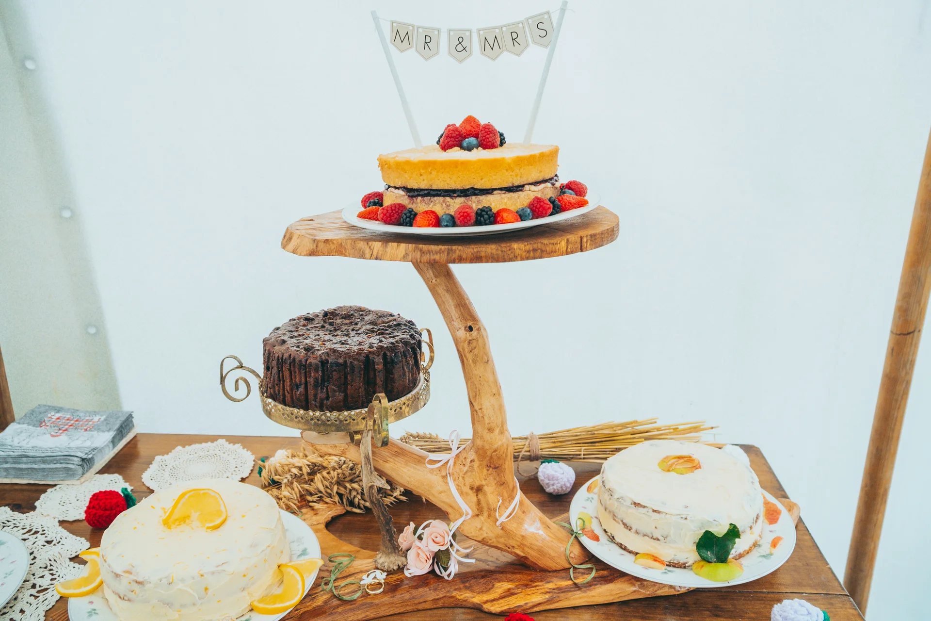 A rustic wedding dessert table featuring a multi-tiered wooden stand holding several delicious cakes topped with fresh berries, citrus slices, and a Mr and Mrs bunting topper.