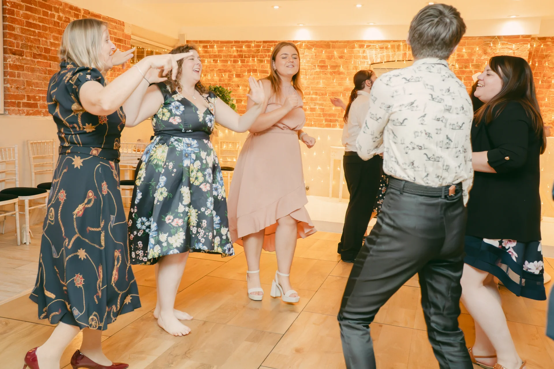 Group of wedding guests laughing and dancing energetically on a wooden dance floor in front of an exposed brick wall decorated with string lights.