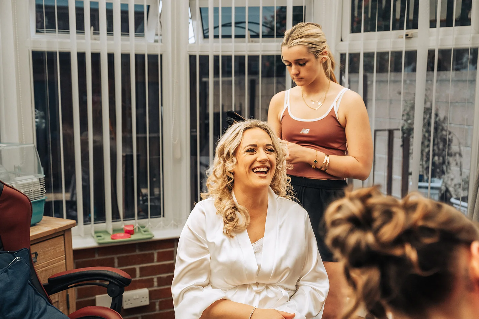 A smiling bride wearing a white satin robe laughs joyfully while having her blonde hair styled by a bridesmaid during morning preparations in a sunlit room.