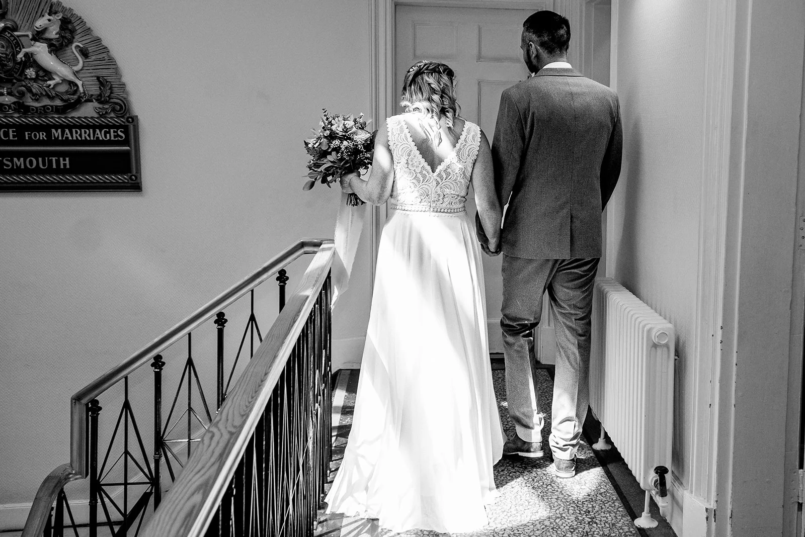 A newlywed bride holding a bouquet and groom walk hand in hand up a staircase inside a registry office, captured in a timeless black and white style.