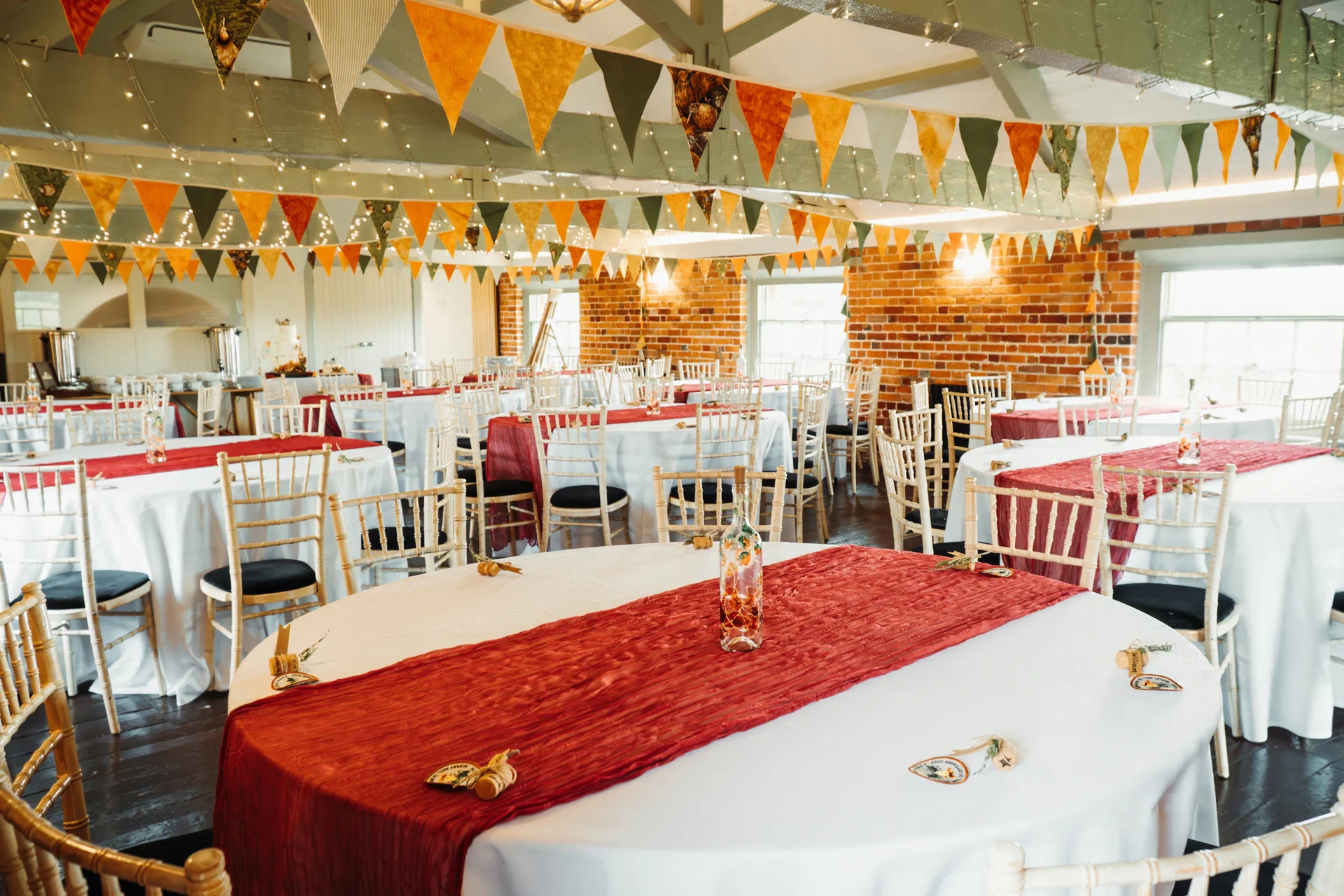 An empty wedding reception venue featuring round tables with white linens and rich red runners, beneath a wooden rafter ceiling decorated with autumn-coloured bunting and fairy lights.