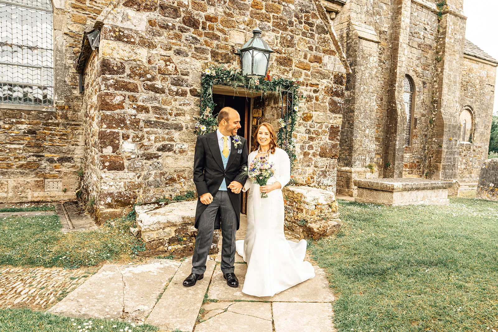 A newlywed bride and groom smiling happily as they step out of an ancient stone church doorway decorated with floral arches.