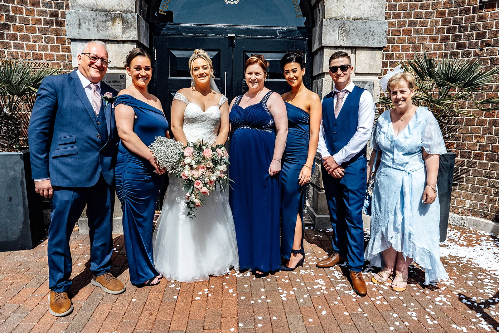 A newlywed couple poses outside a historic brick registry office with their family and bridal party under bright sunlight on the pavement.