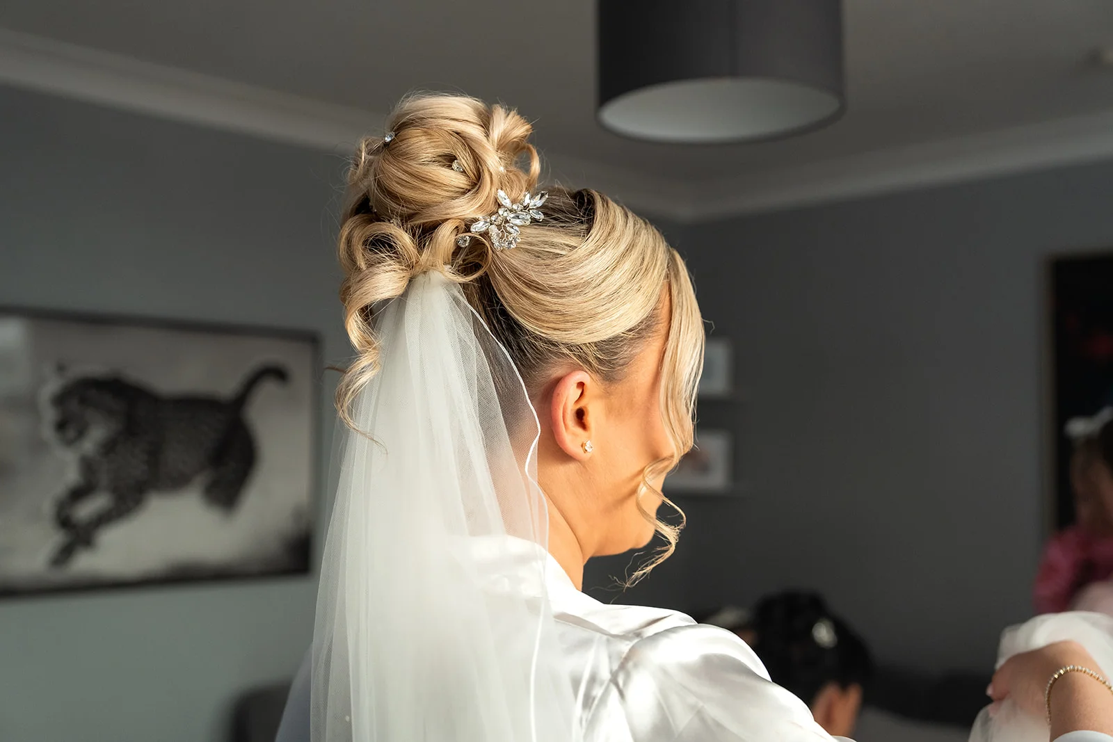 A close-up back view of a blonde bride during bridal preparations, showcasing an intricate high bun adorned with a crystal hair comb and a delicate veil.