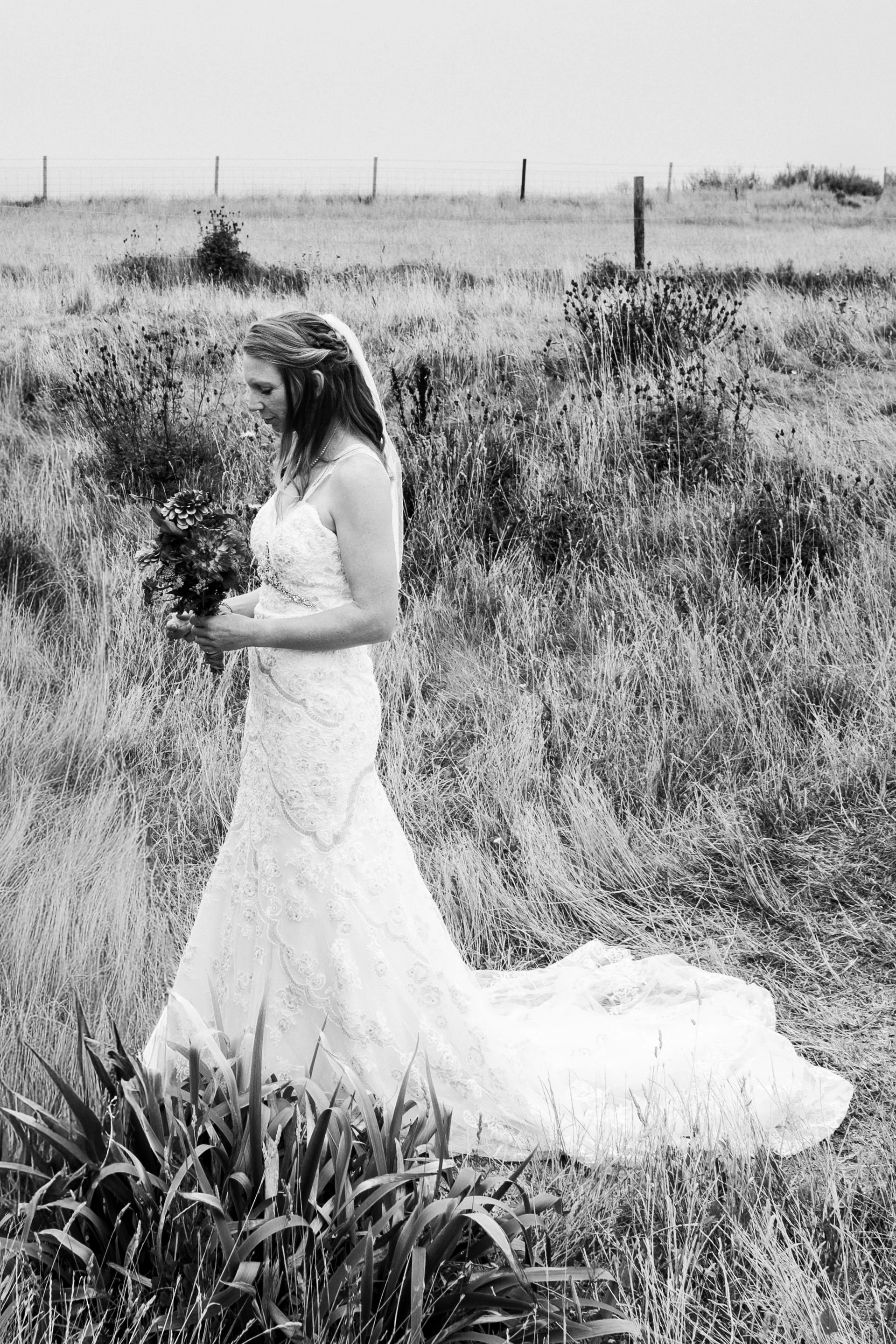 A black and white portrait of a bride standing in a rural grassy field holding a bouquet while wearing a lace fit and flare wedding dress with a train.