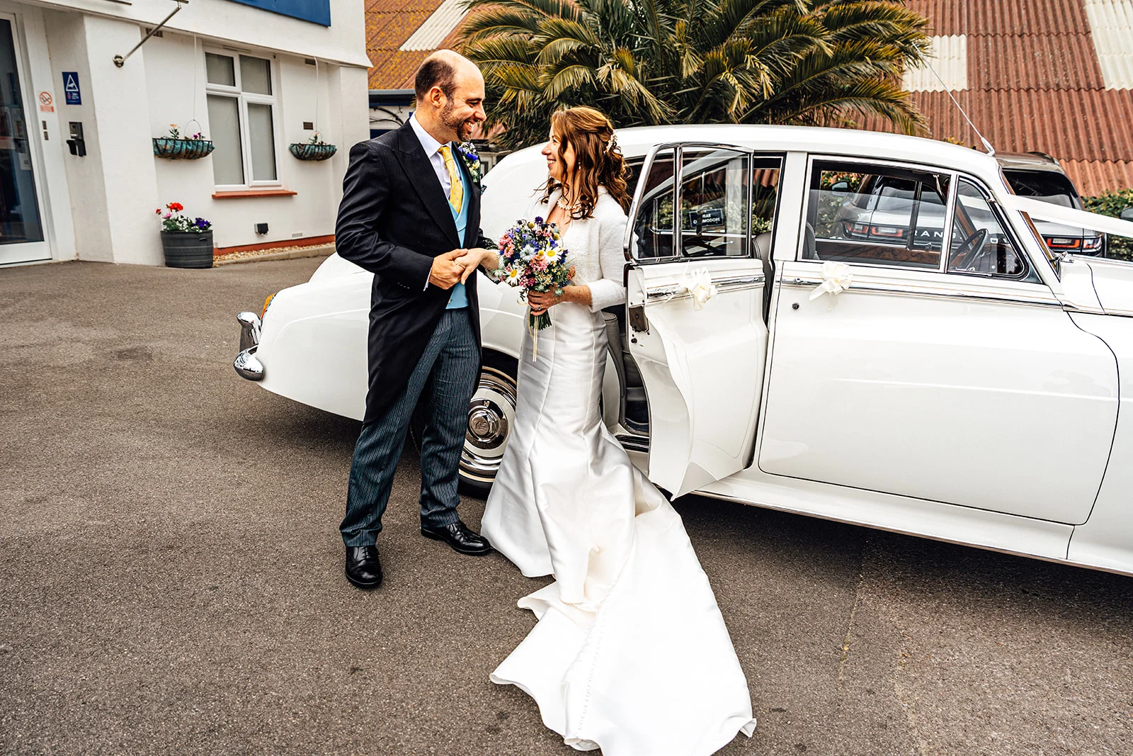 A smiling bride and groom holding hands next to a white vintage wedding car outside a building.