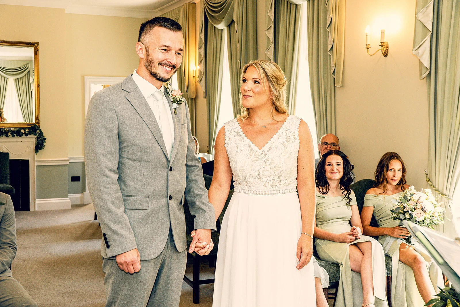 A smiling groom in a grey suit and a bride in a lace-bodice gown hold hands during their light-filled civil ceremony indoors.
